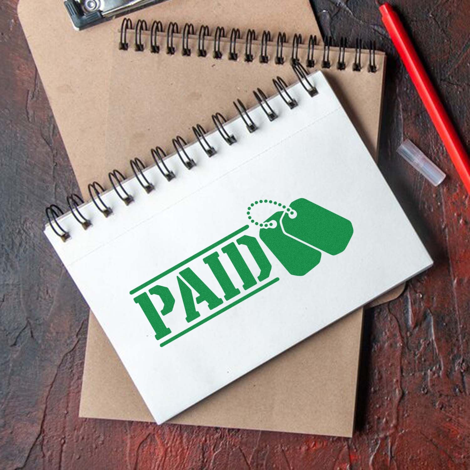 White spiral notebook with green PAID stamp and seal graphic, on kraft paper beside a red pen.