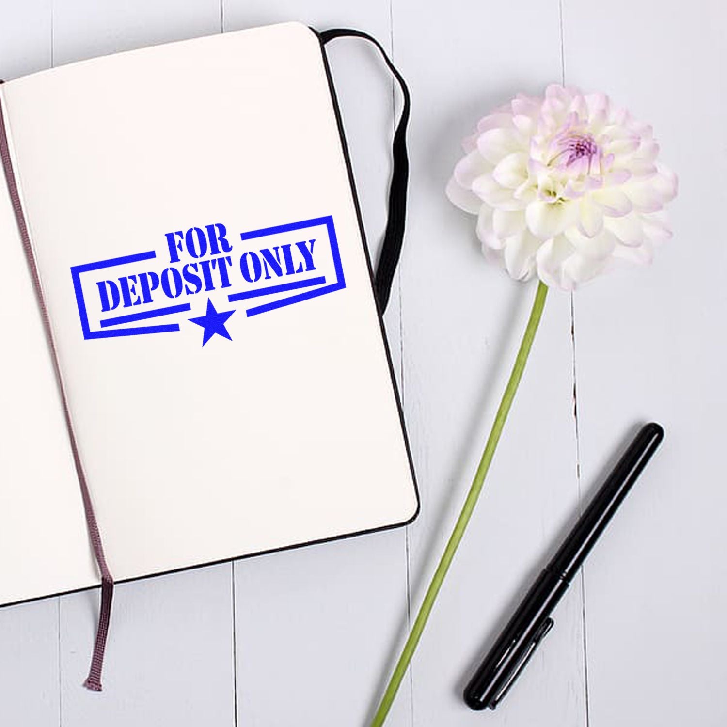 White notebook stamped with a blue For Deposit Only seal, beside a black pen and white flower on a white wood desk