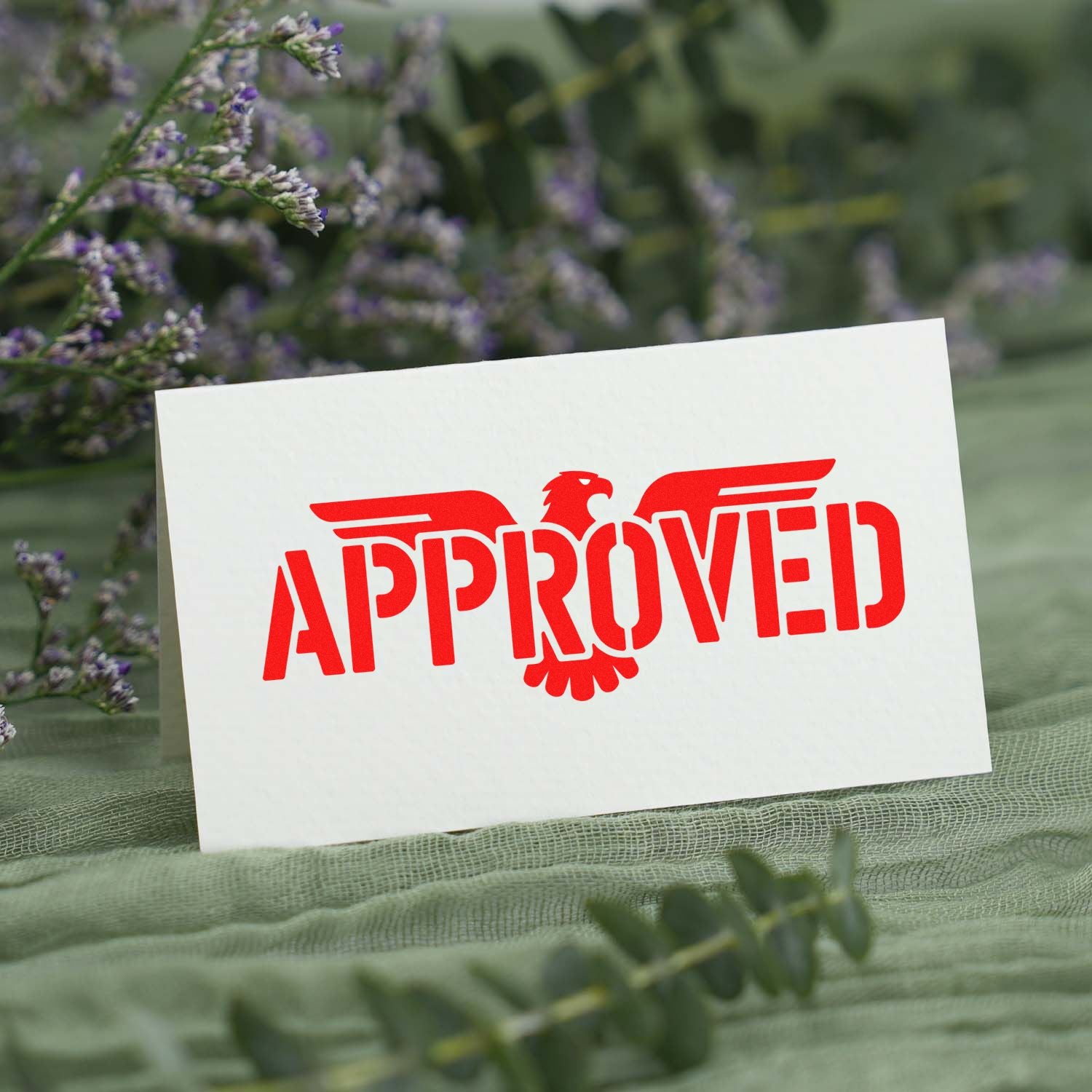 Red APPROVED eagle seal stamp on a white card, set against green foliage and purple flowers.
