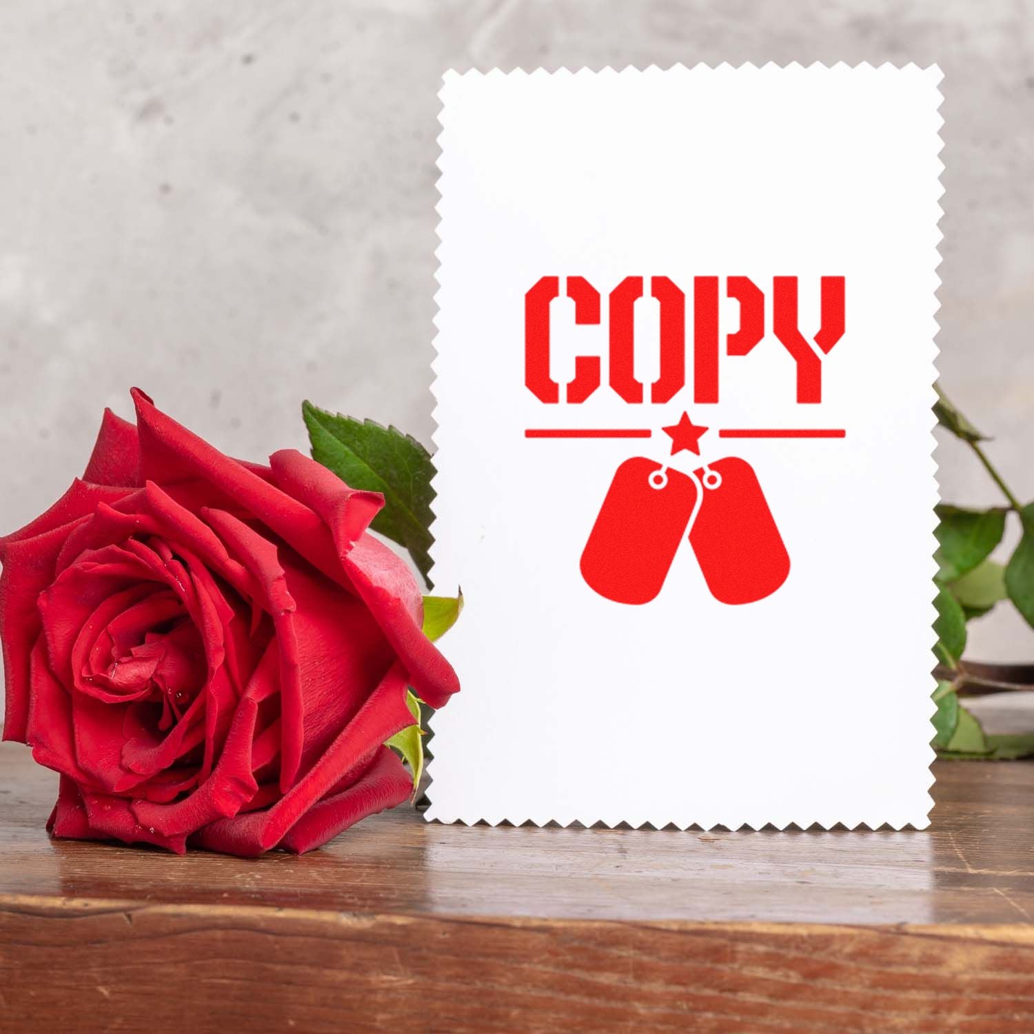 Red rose beside a white stamp-and-seal card with red COPY text and dog tag emblem on a wooden table.