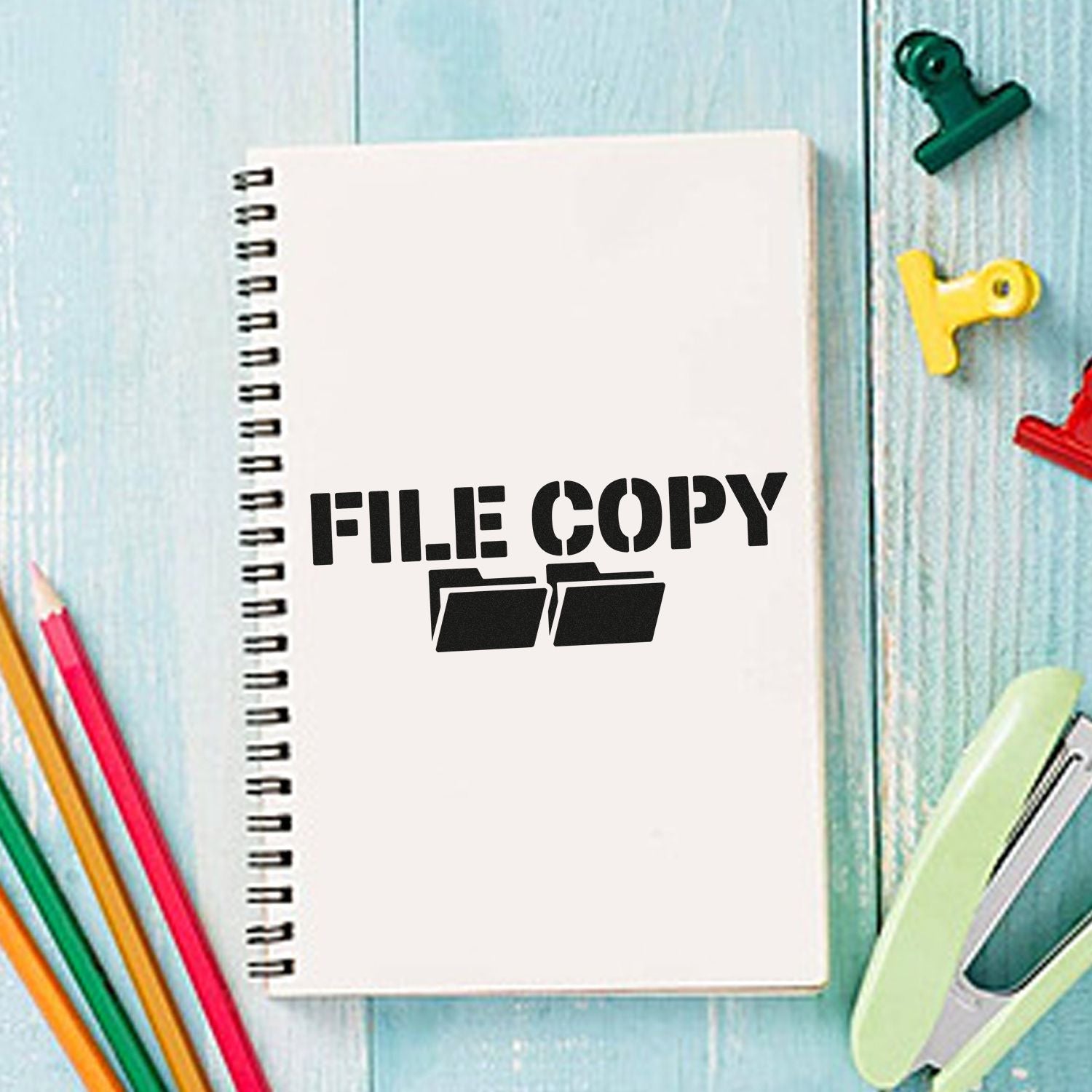 White spiral notebook with black FILE COPY stamp seal, surrounded by colorful pencils, binder clips, and paper clips.