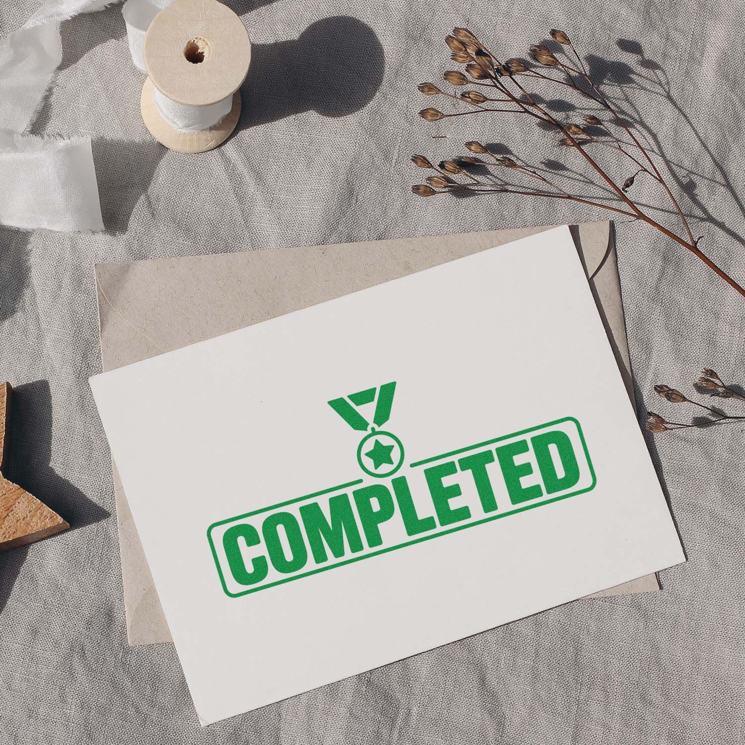 White card with green COMPLETED stamp and medal seal over beige envelope, with ribbon spool and dried stems.