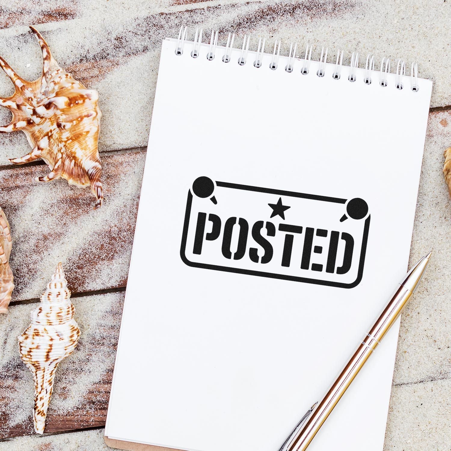 White spiral notepad with black POSTED stamp seal, seashells, and gold pen on a sandy beach background.