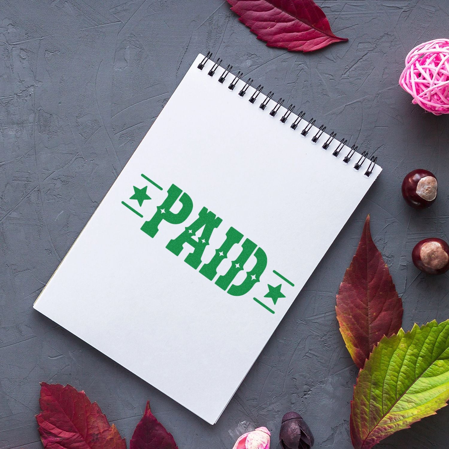 White spiral notebook with green PAID stamp seal on gray background, surrounded by burgundy and green leaves.
