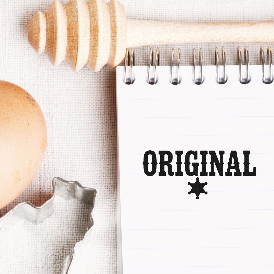 Notebook stamped ORIGINAL with black seal badge, beside a wooden honey dipper, egg, and metal cookie cutter on beige cloth.