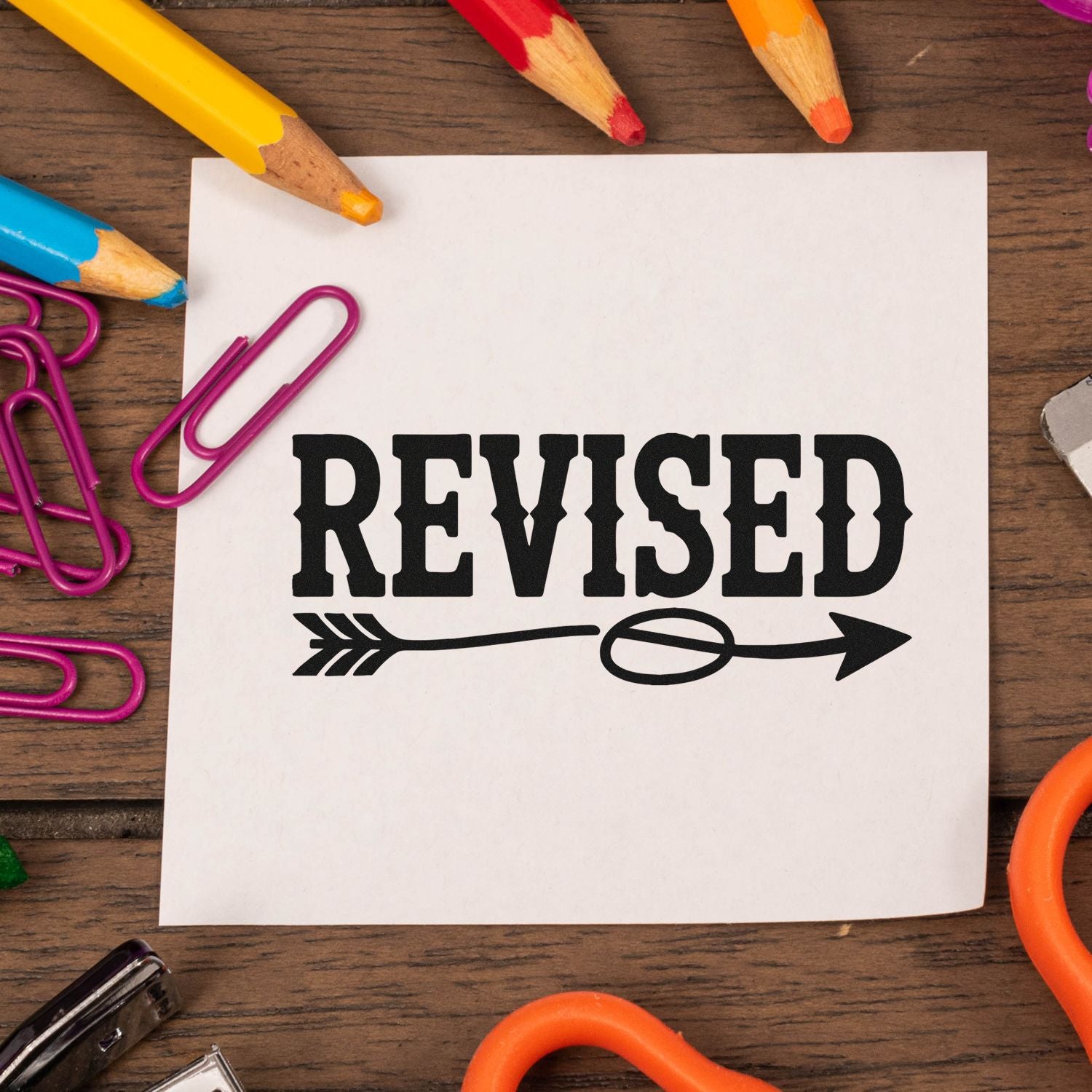 Black REVISED stamp seal on white paper, surrounded by colorful pencils and magenta paper clips.