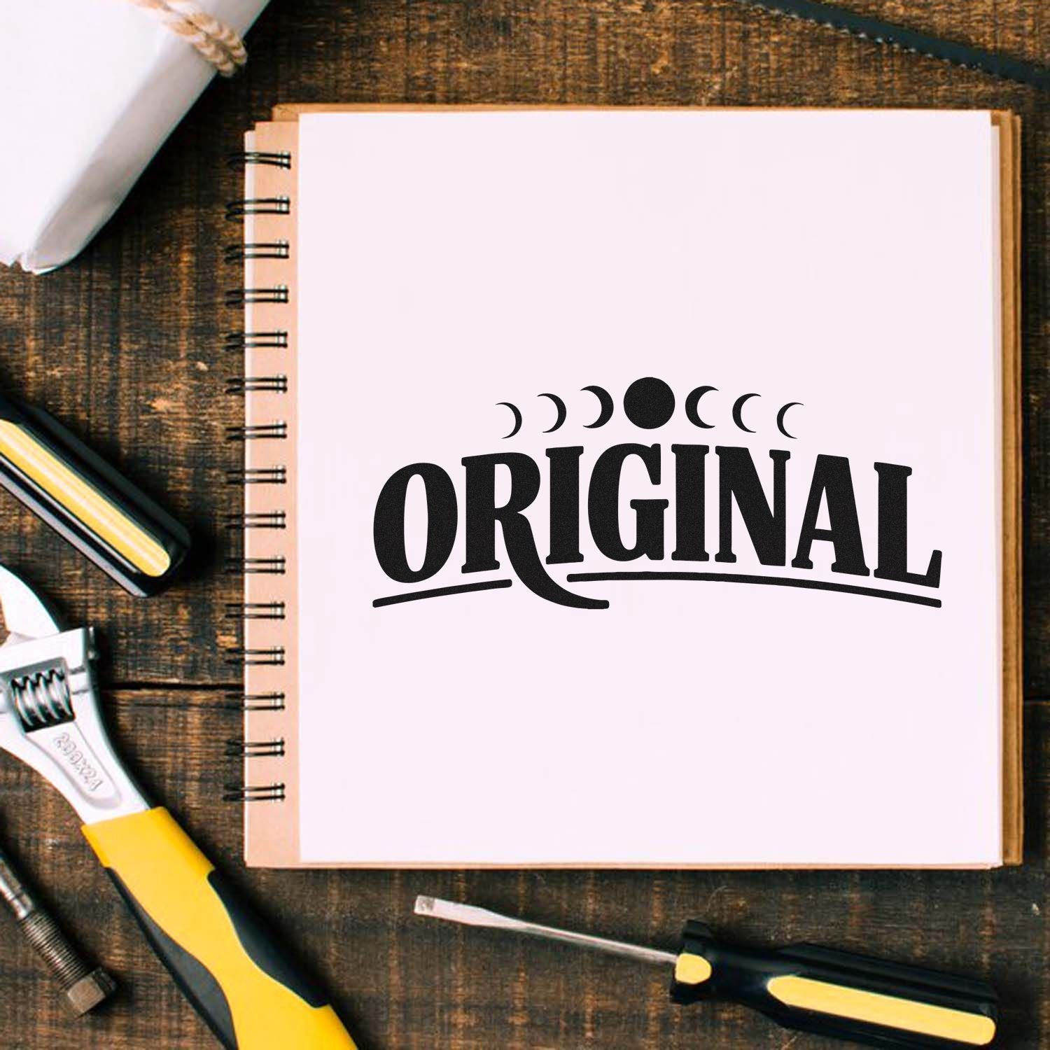 Black ORIGINAL seal stamp with moon phases on a white spiral notebook, surrounded by yellow-and-black tools on dark wood.