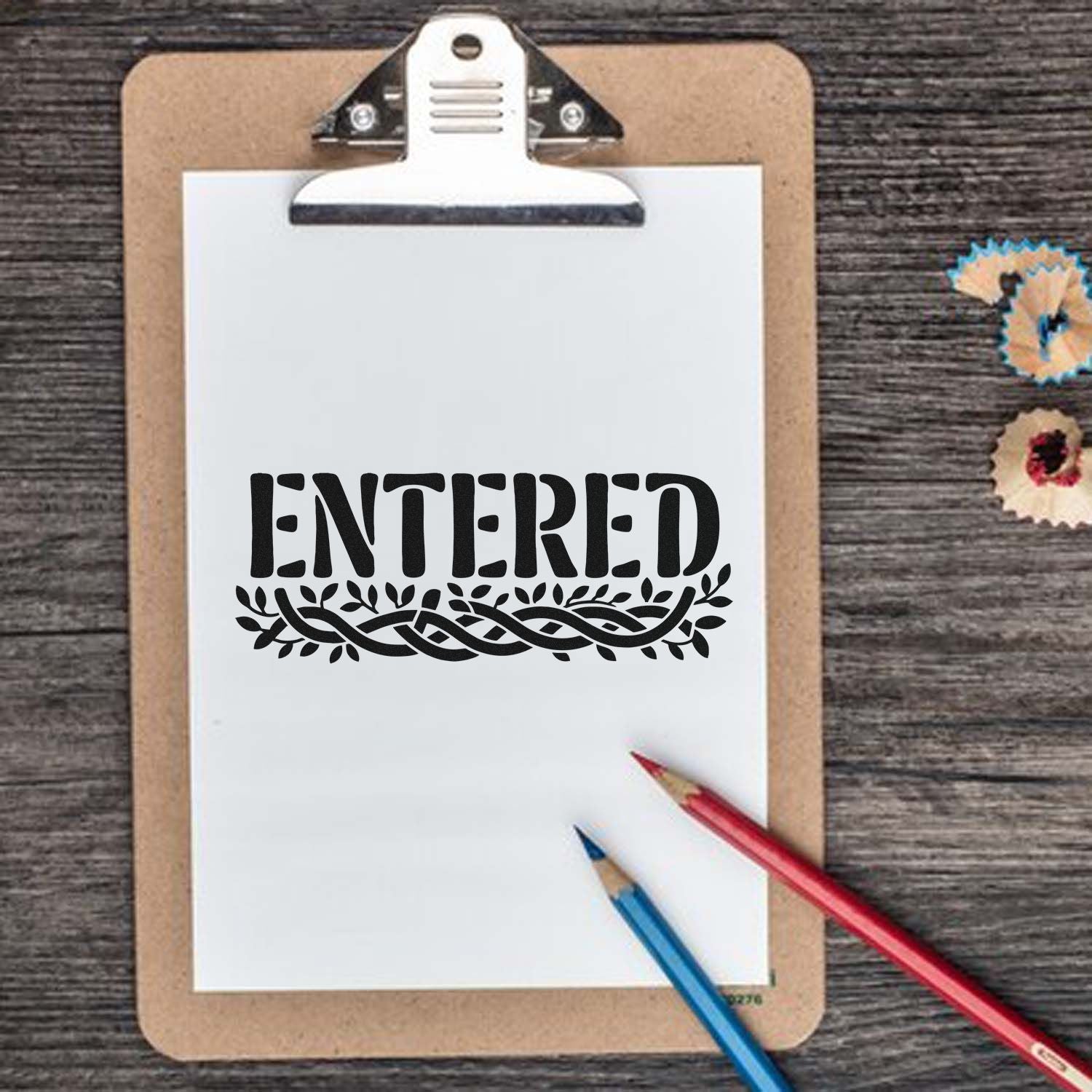Black ENTERED stamp on white clipboard with leaf border, tan board, red and blue pencils, and decorative seal samples.