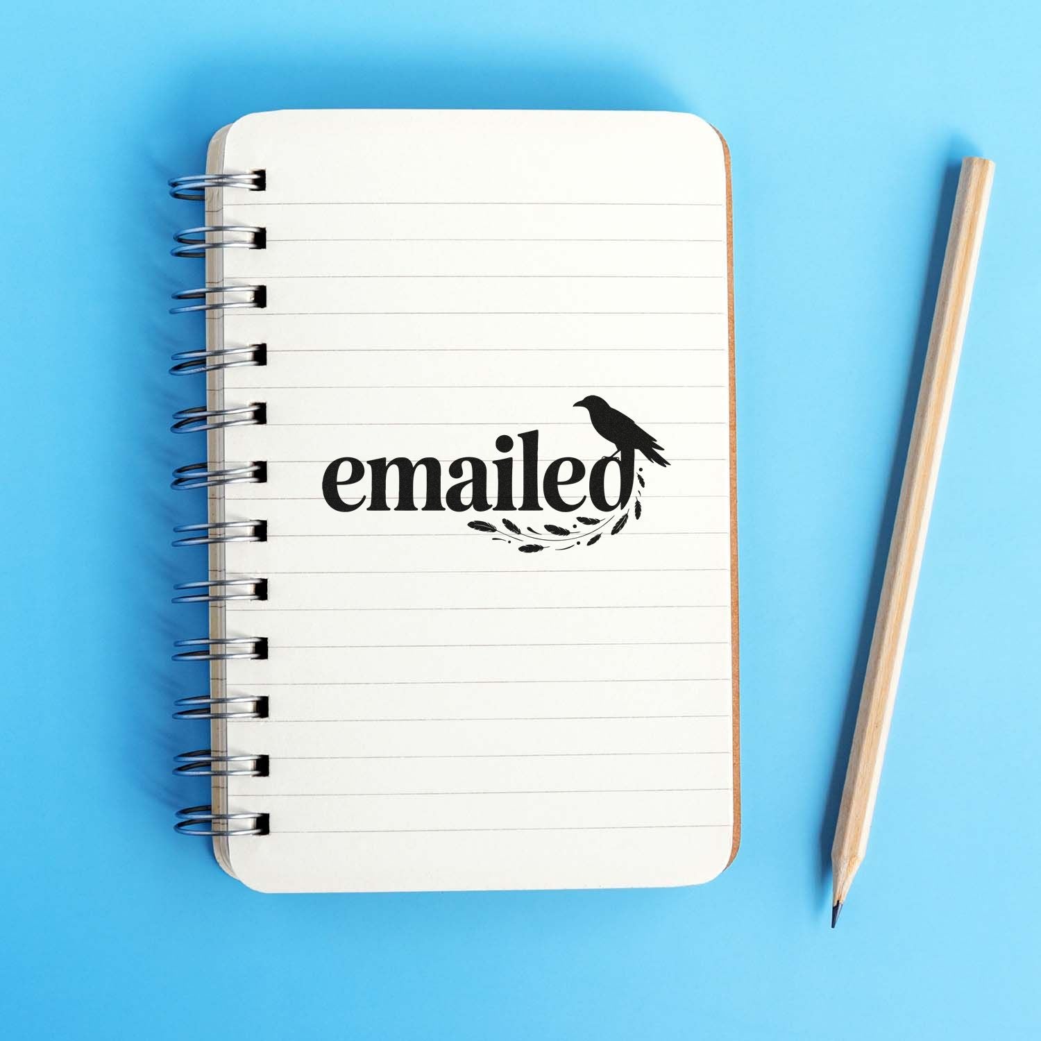 White spiral notebook with black emailed stamp and bird seal on a blue background, beside a wooden pencil.