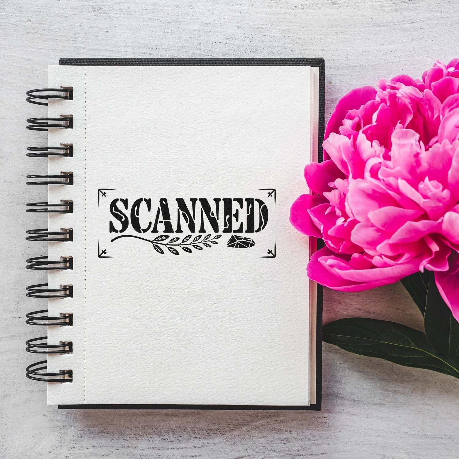 Black SCANNED stamp seal on white spiral notebook, beside bright pink peonies and a dark cover