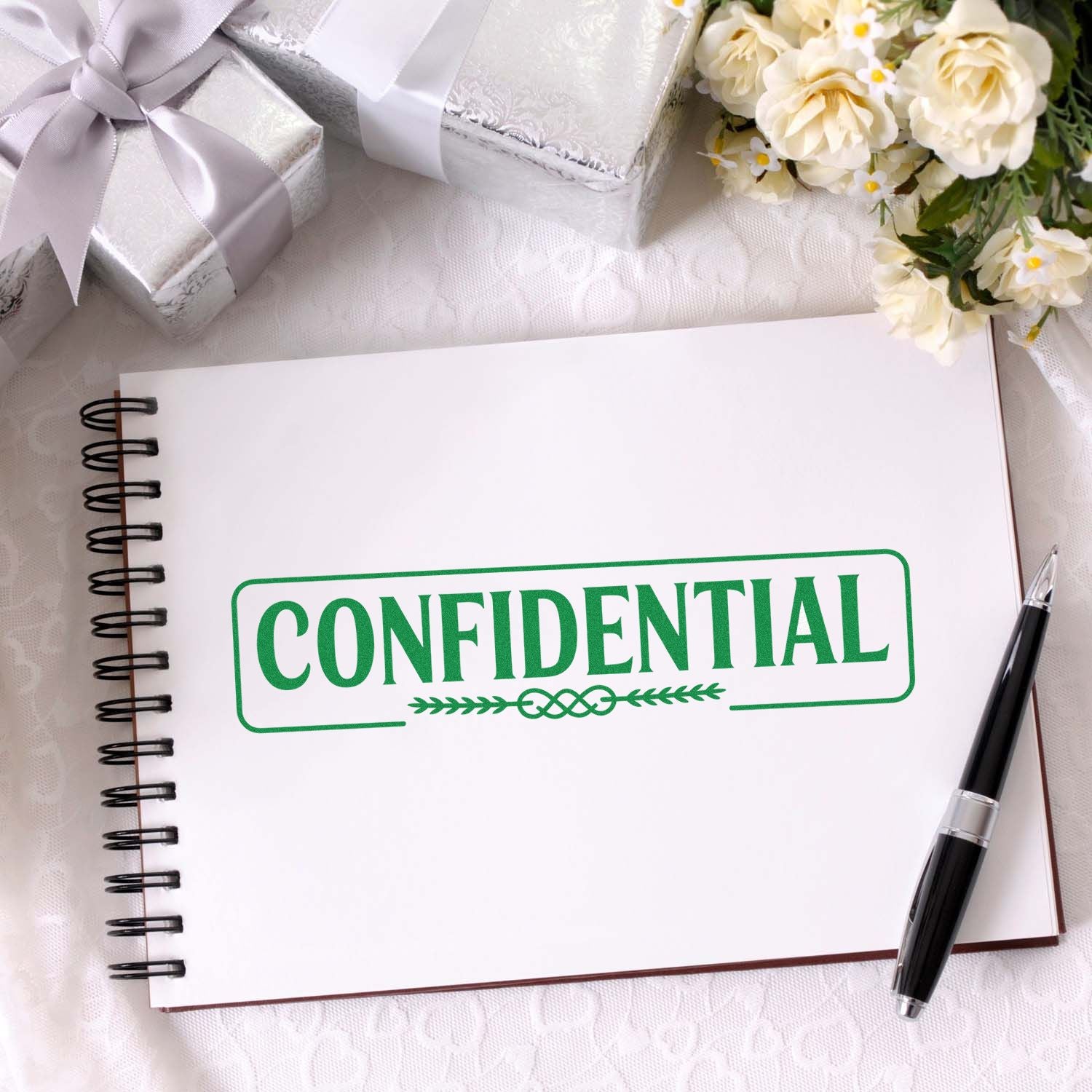 Green confidential stamp seal on a white spiral notebook, with silver gift boxes, cream flowers, and a black pen.