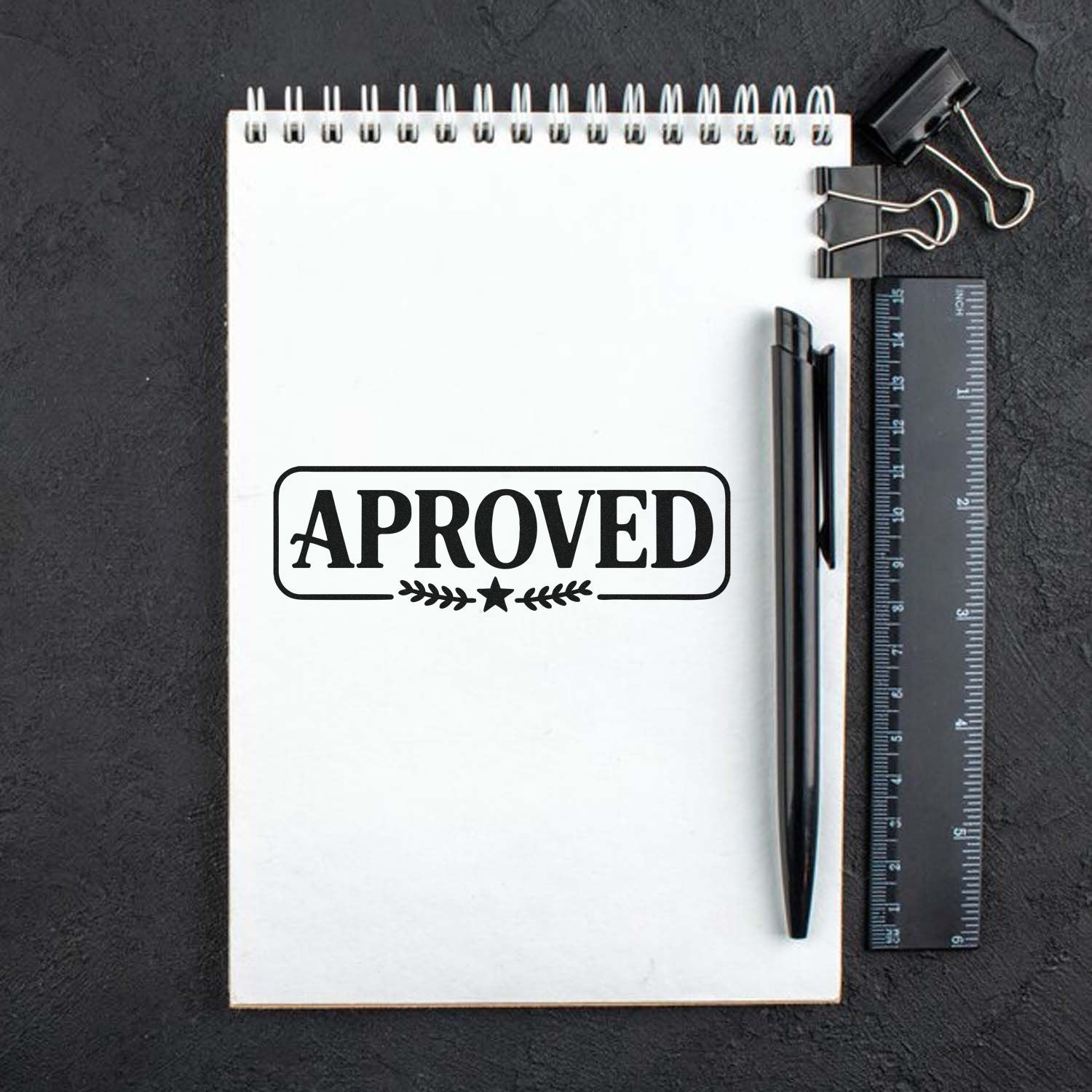 Black APPROVED rubber stamp seal on white spiral notebook with black pen, ruler, and binder clip on dark background.