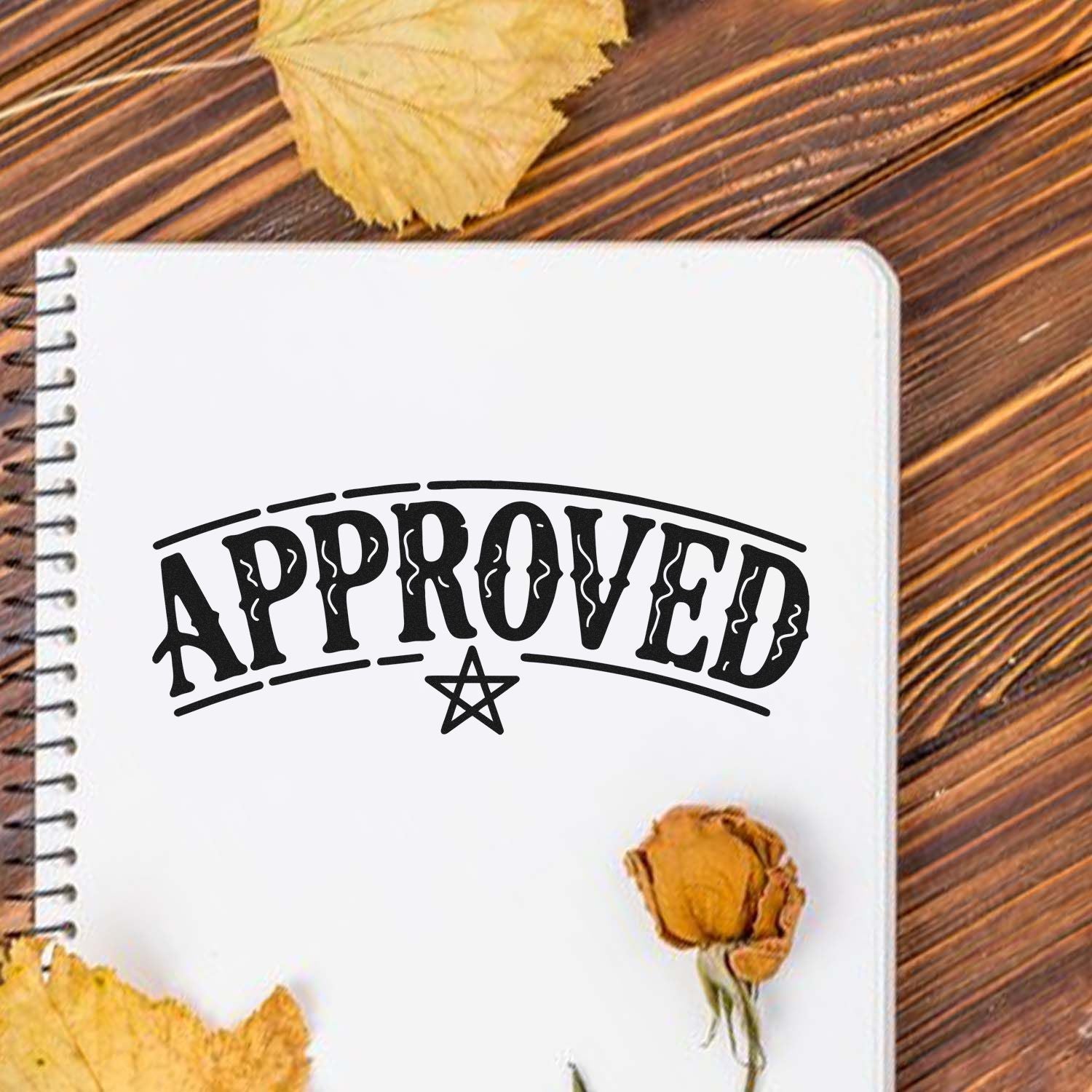 Black APPROVED rubber stamp seal with star on a white notebook, styled on rustic wood with dried flowers.