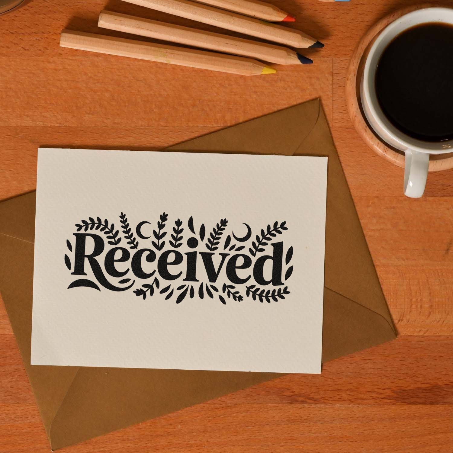 Black floral Received stamp on white card, with brown envelope, coffee cup, and colored pencils on a wooden desk.