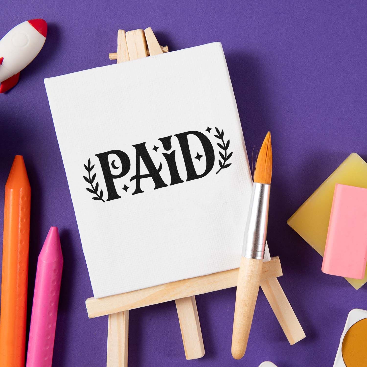 Black PAID stamp seal on white canvas easel, surrounded by colorful art tools on a purple background