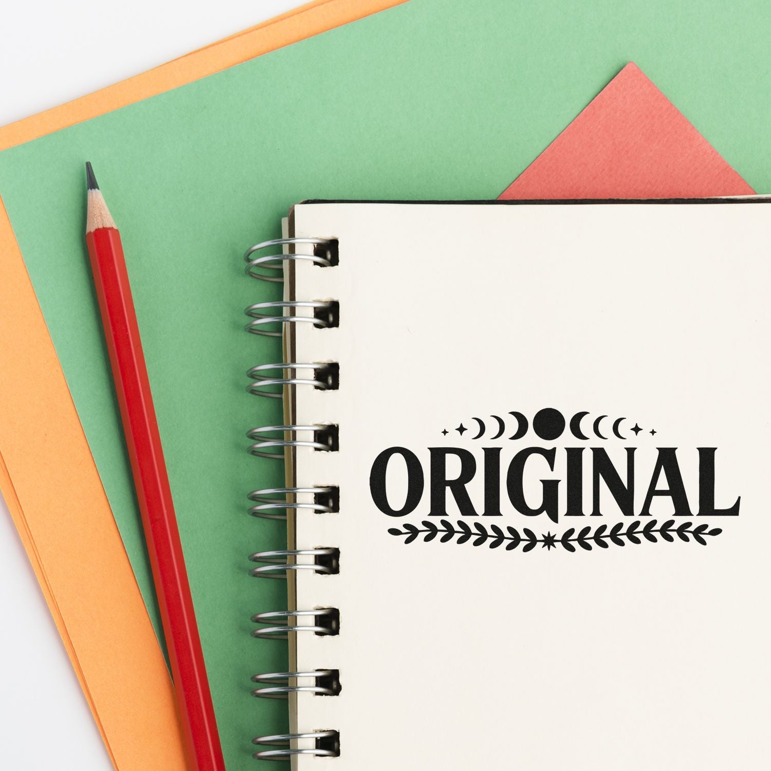 Original stamp and seal design on a cream spiral notebook, with a red pencil and green-orange paper background.