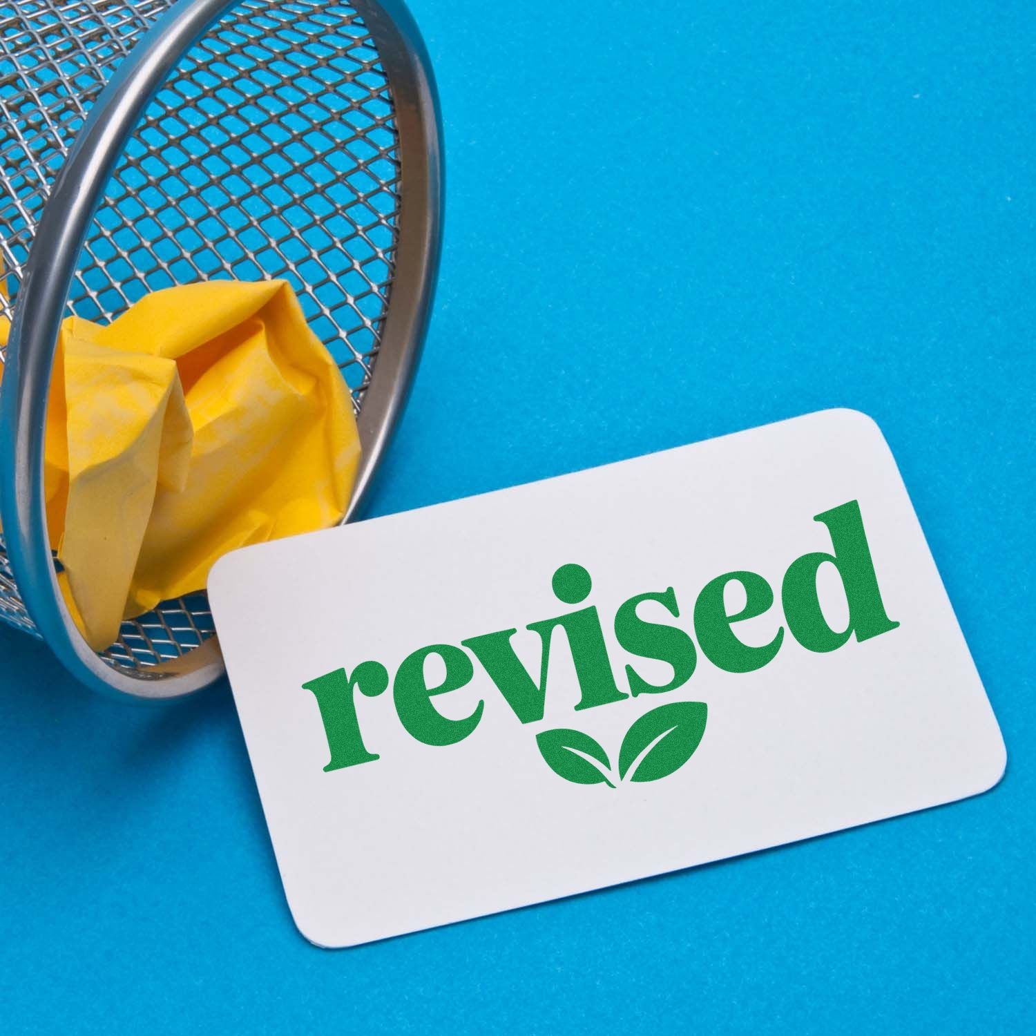 Green revised stamp and seal card with leaf icon beside a metal mesh basket and yellow paper on bright blue background.