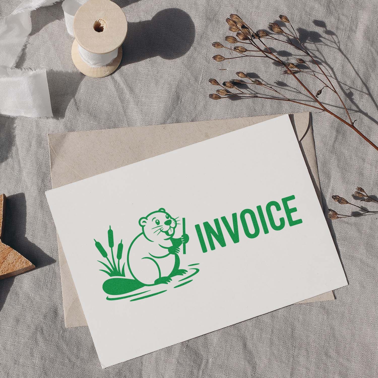 Green custom invoice stamp seal with otter on white card, beside beige envelope, ribbon spool, and dried stems.