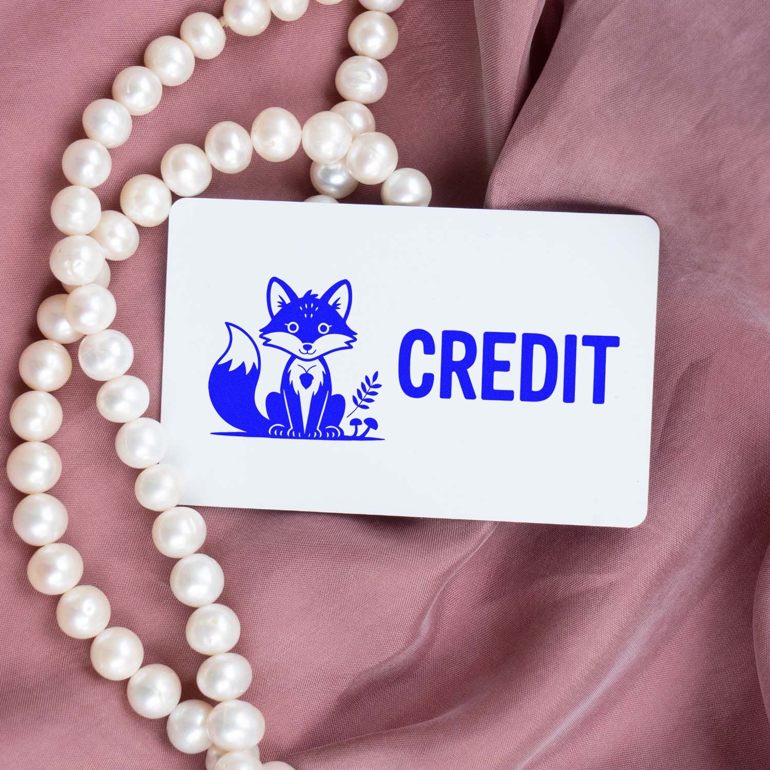 Blue fox CREDIT stamp and seal on white card, resting on pink fabric with white pearl beads.