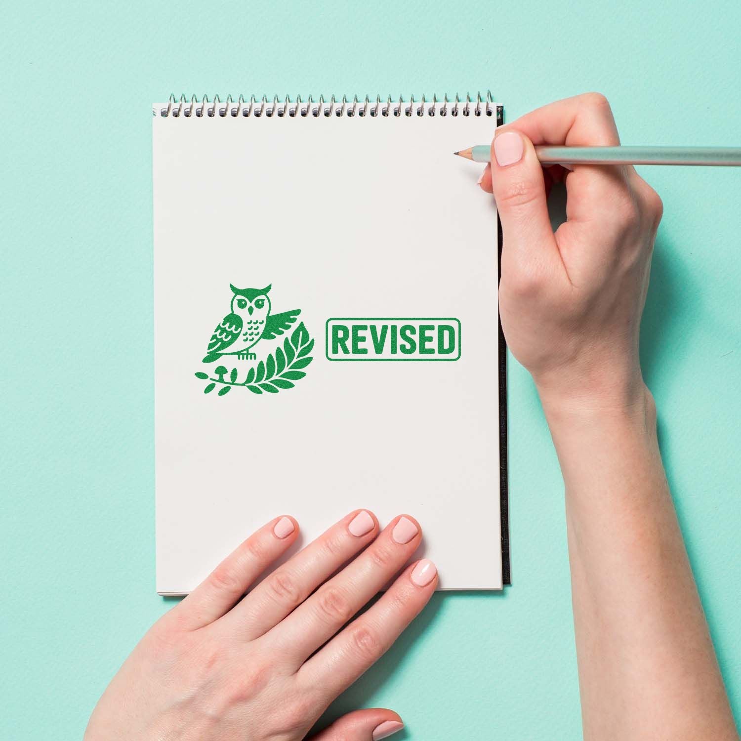 Green revised stamp and owl seal on a white spiral notebook, held by hands on a mint background.