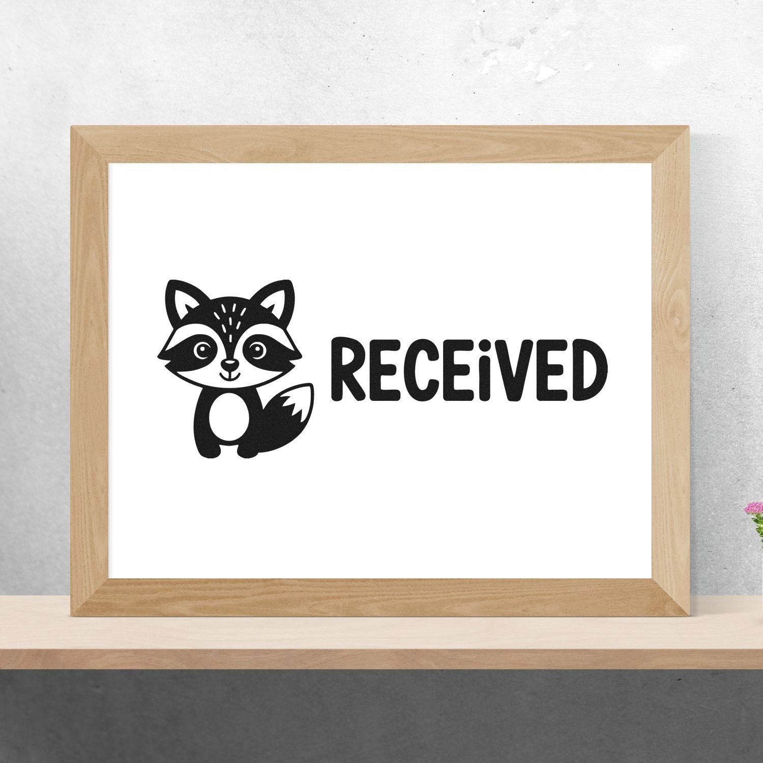 Black raccoon RECEIVED stamp/seal design in black on white, displayed in a light wood frame.