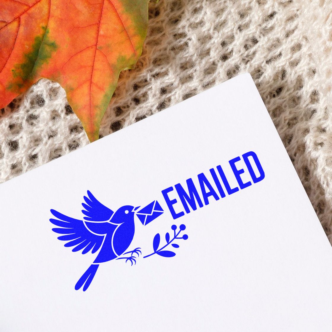 Blue emailed stamp seal with bird carrying envelope, on white paper with autumn leaf and beige knit background.