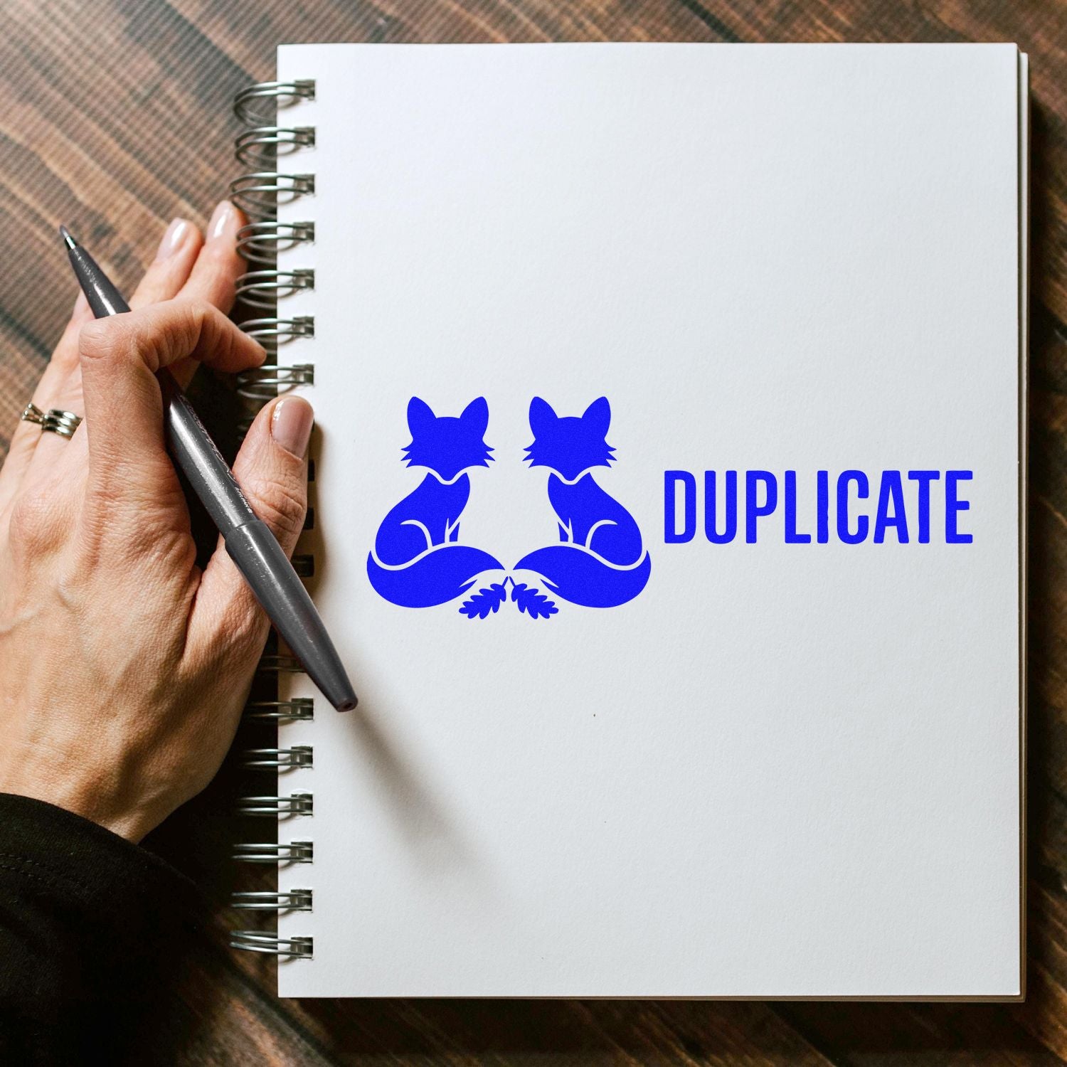 Blue duplicate stamp seal with mirrored fox design on white spiral notebook, held by hand with pen.
