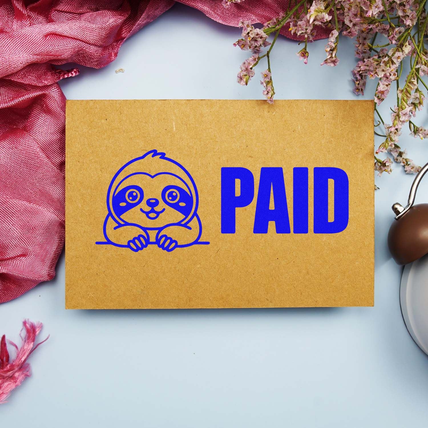 Kraft envelope with blue sloth PAID stamp seal, pink fabric and flowers on a light blue background.