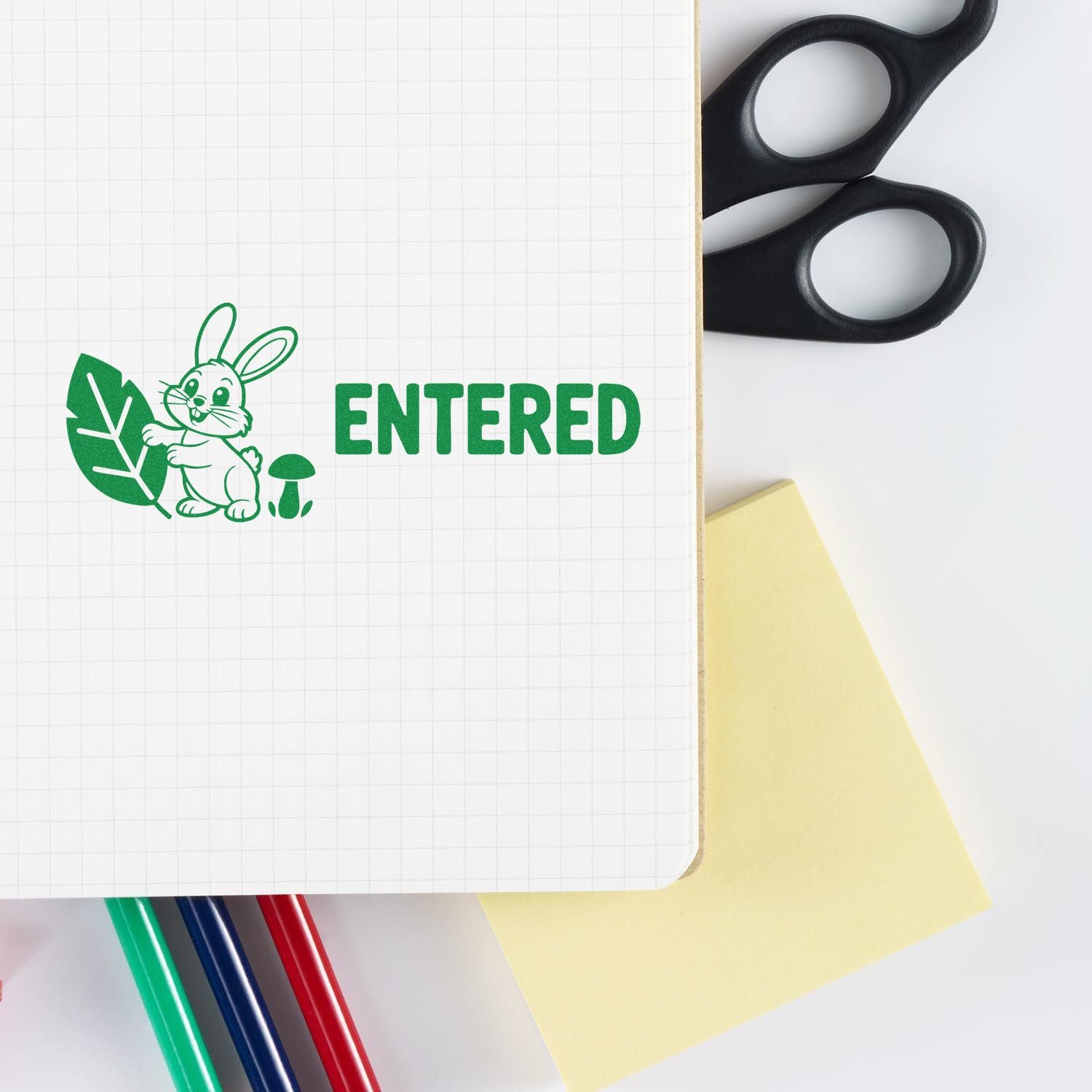 Green ENTERED stamp with bunny, leaf and mushroom seal on notebook, beside black scissors, yellow sticky note and colored pens.