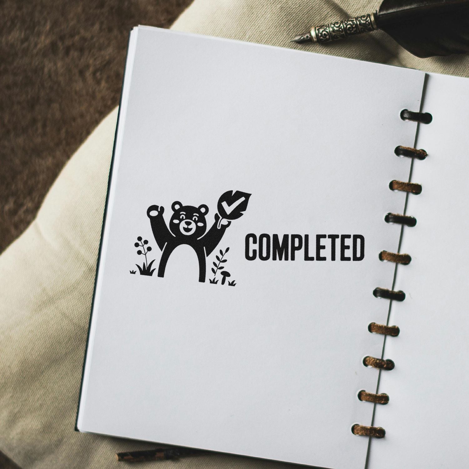 Black completed stamp seal with bear and checkmark on white notebook, beside metal spiral binding.