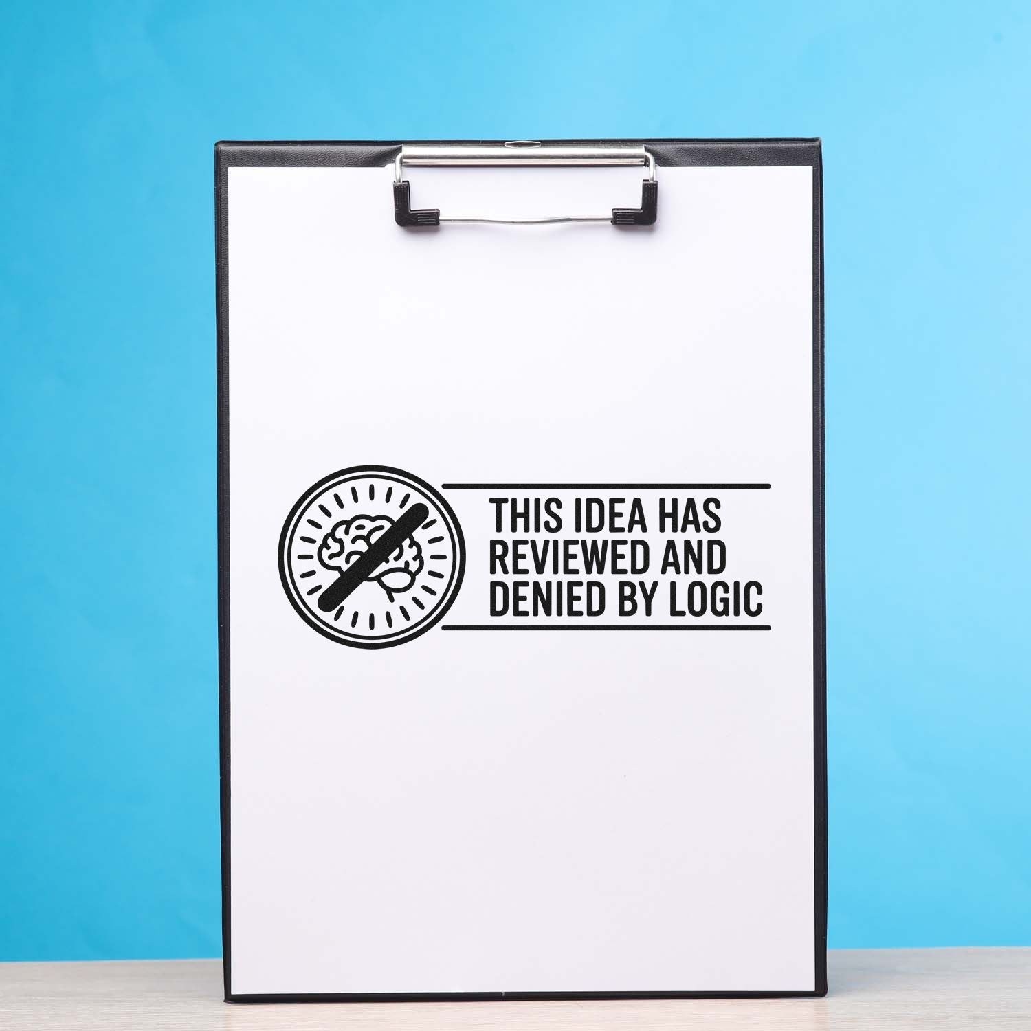 Black stamp seal on a white clipboard reading "This idea has reviewed and denied by logic" with a crossed-out brain, blue background.