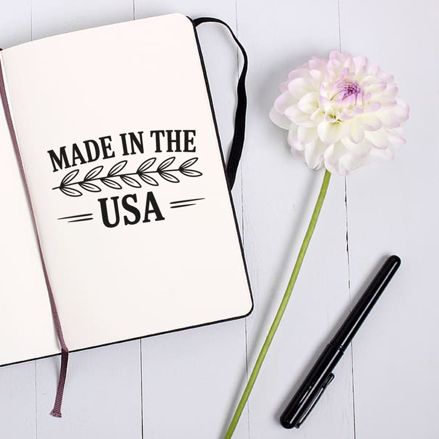 Notebook with black Made in the USA seal stamp, black pen, and white flower on a light wood background.