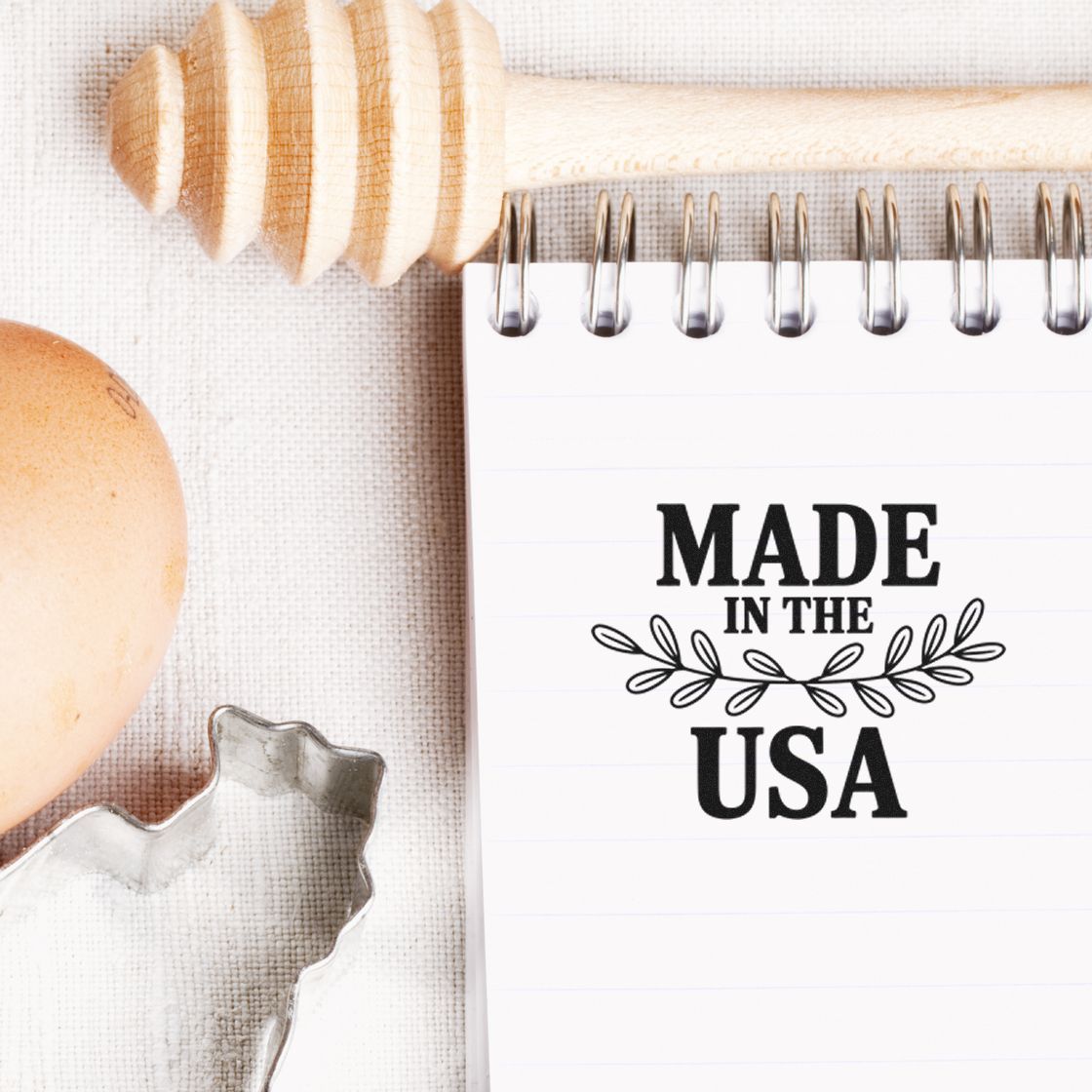 Black Made in USA stamp with laurel on white notepad, beside wooden honey dipper, egg, and metal cookie cutter.