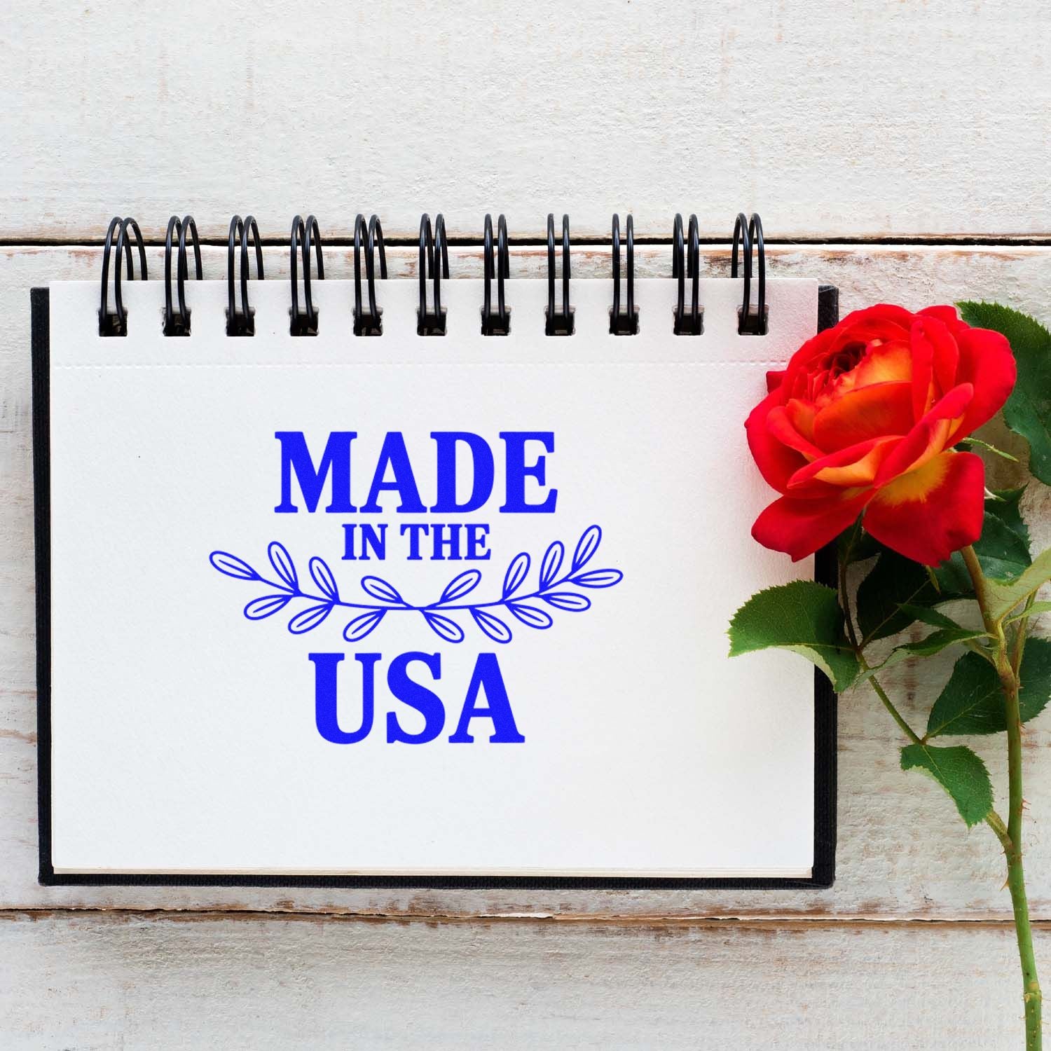 Blue Made in USA stamp seal on white spiral notebook with black binding, beside a red rose.
