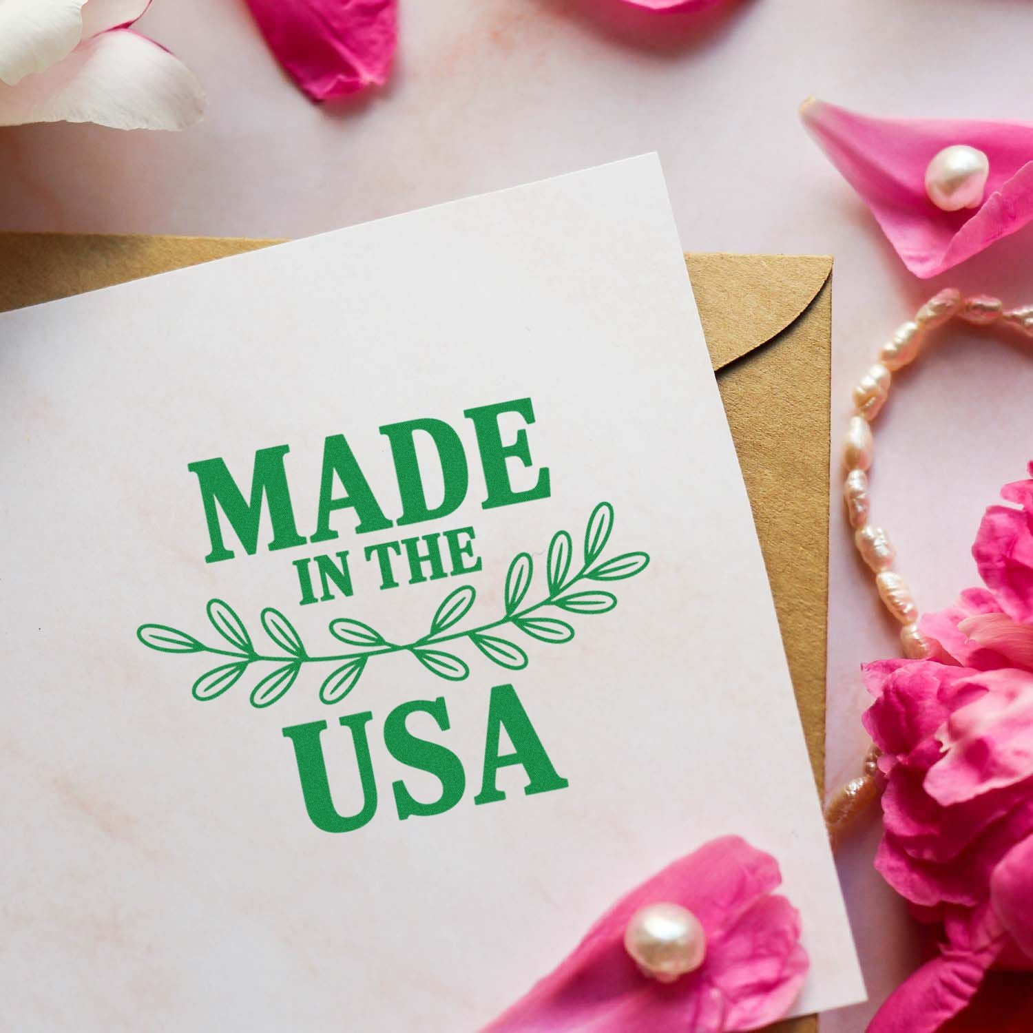 Green Made in the USA stamp and seal on white card over tan envelope, with pink petals and pearl accents.