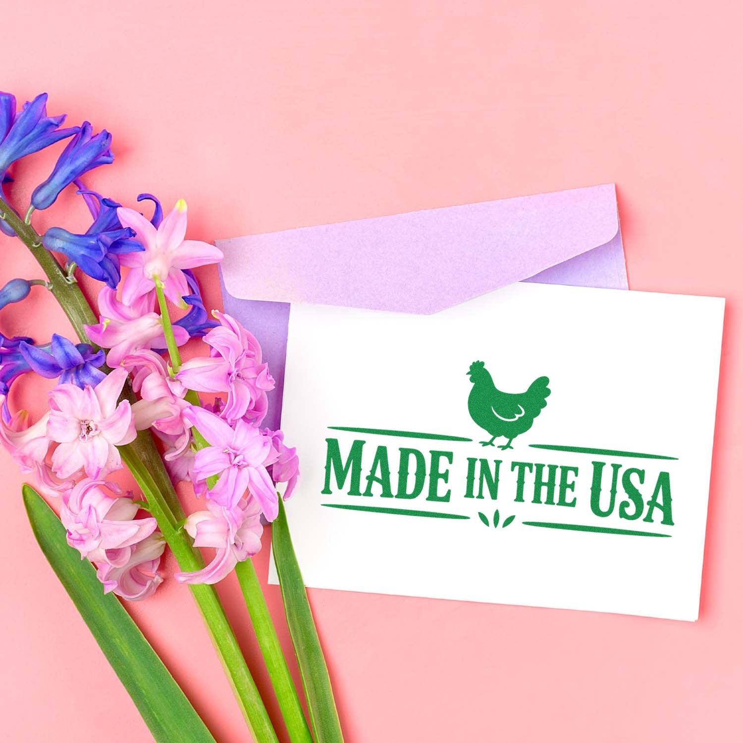 Green Made in the USA stamp seal on white card with purple envelope and pink-purple flowers on a soft pink background.