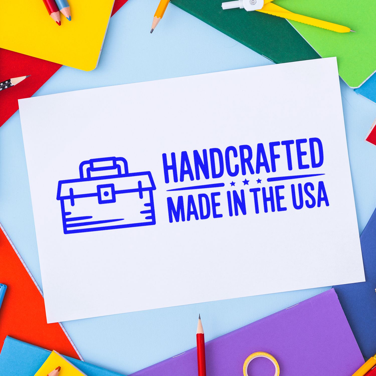 Blue handcrafted Made in the USA rubber stamp seal on white paper, surrounded by colorful pencils and notebooks.