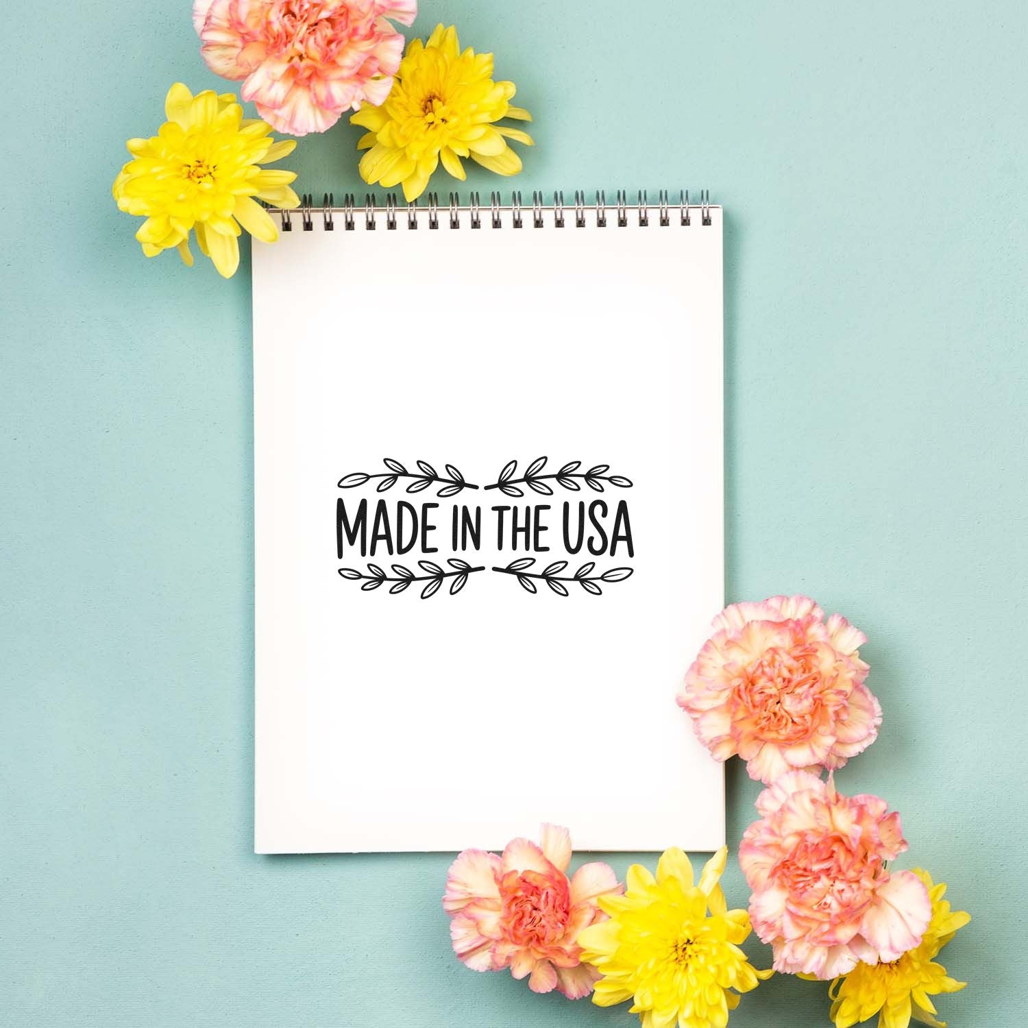White spiral notebook page with black Made in the USA stamp seal, framed by yellow and pink flowers on a mint background.