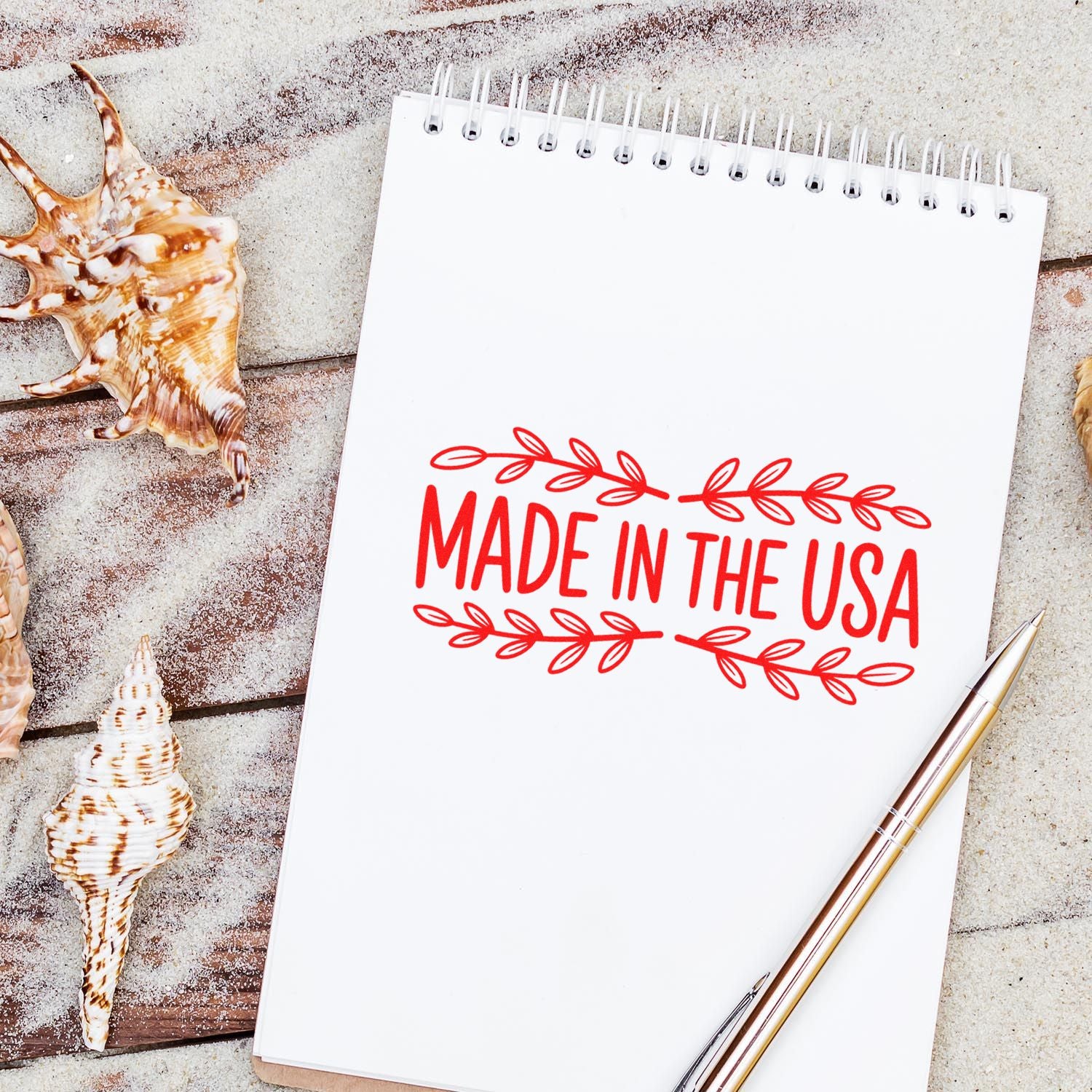 Red Made in the USA stamp and seal on a white spiral notepad, with seashells and a gold pen on a sandy background.