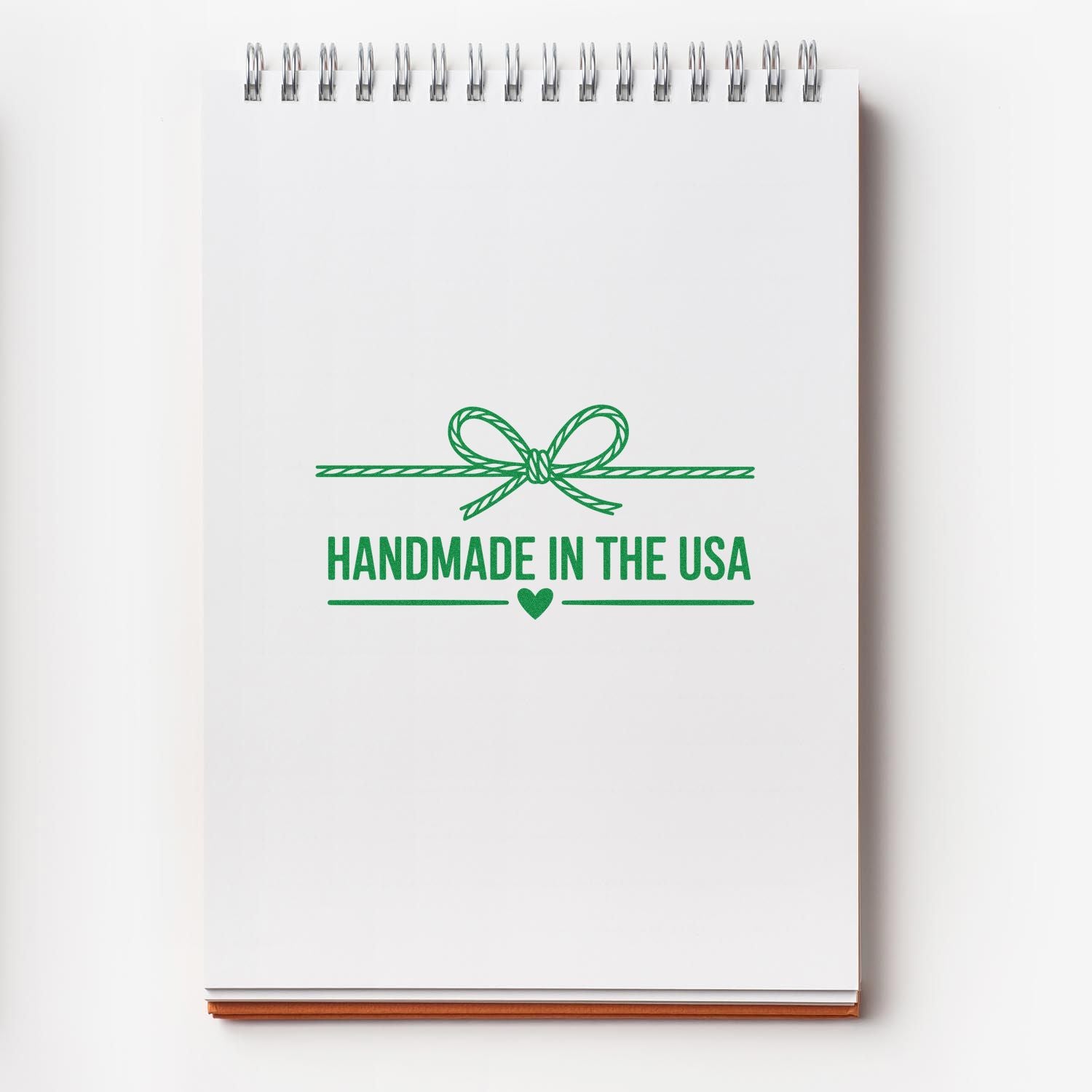 White spiral notebook with green Handmade in the USA stamp seal, ribbon bow and heart emblem.