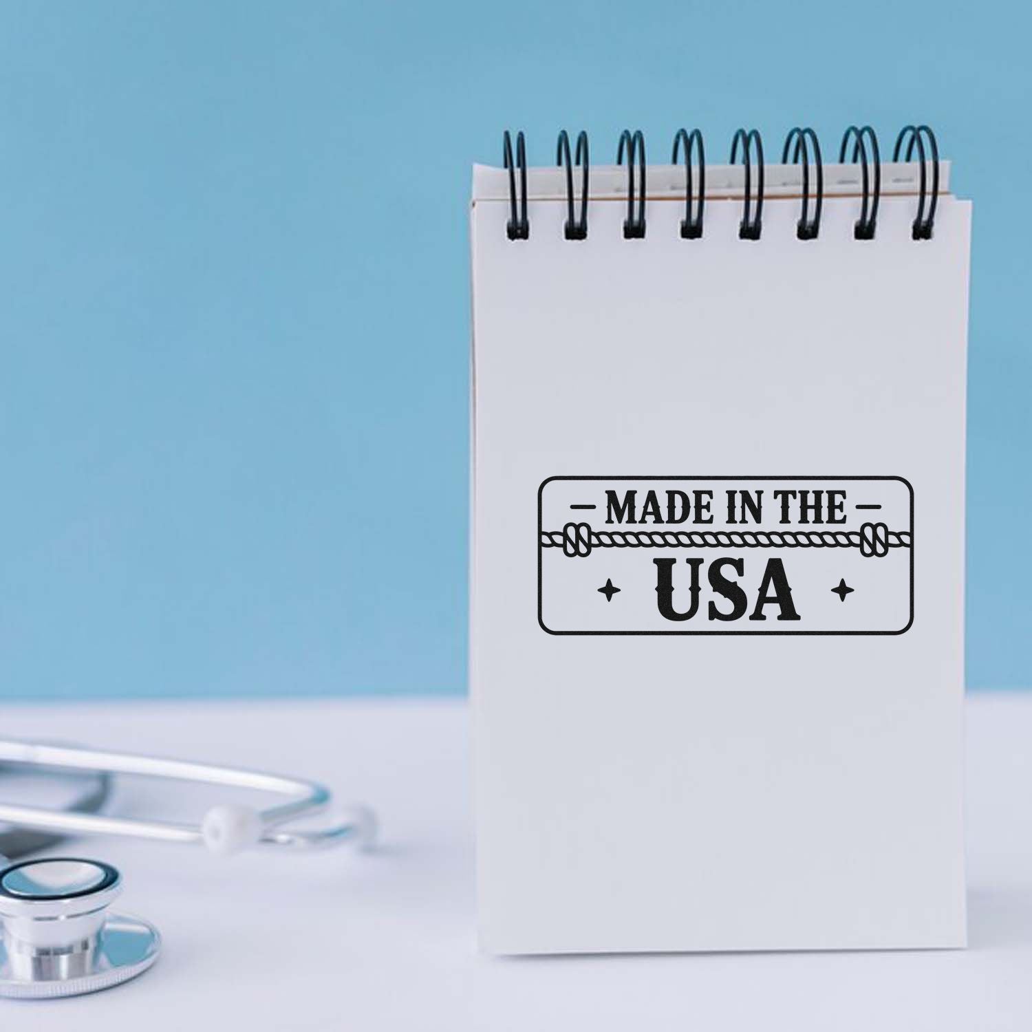 White spiral notepad with black Made in the USA stamp seal on a light blue background, with a stethoscope at the bottom.