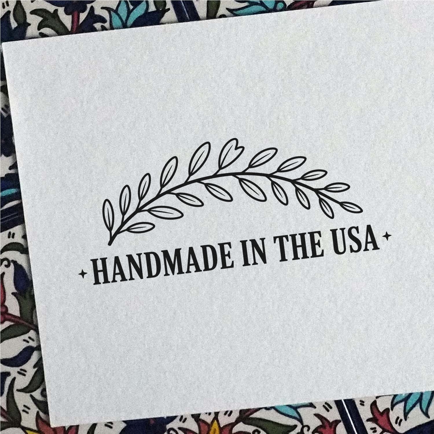 Black handmade-in-the-USA stamp seal with leafy wreath on white paper over colorful patterned fabric.