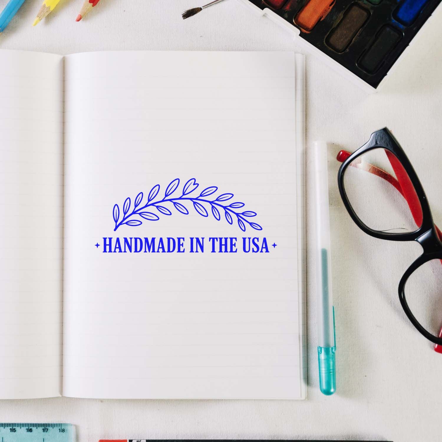 Blue Handmade in the USA seal stamp with leafy wreath on open notebook, beside glasses, pens, and art supplies.