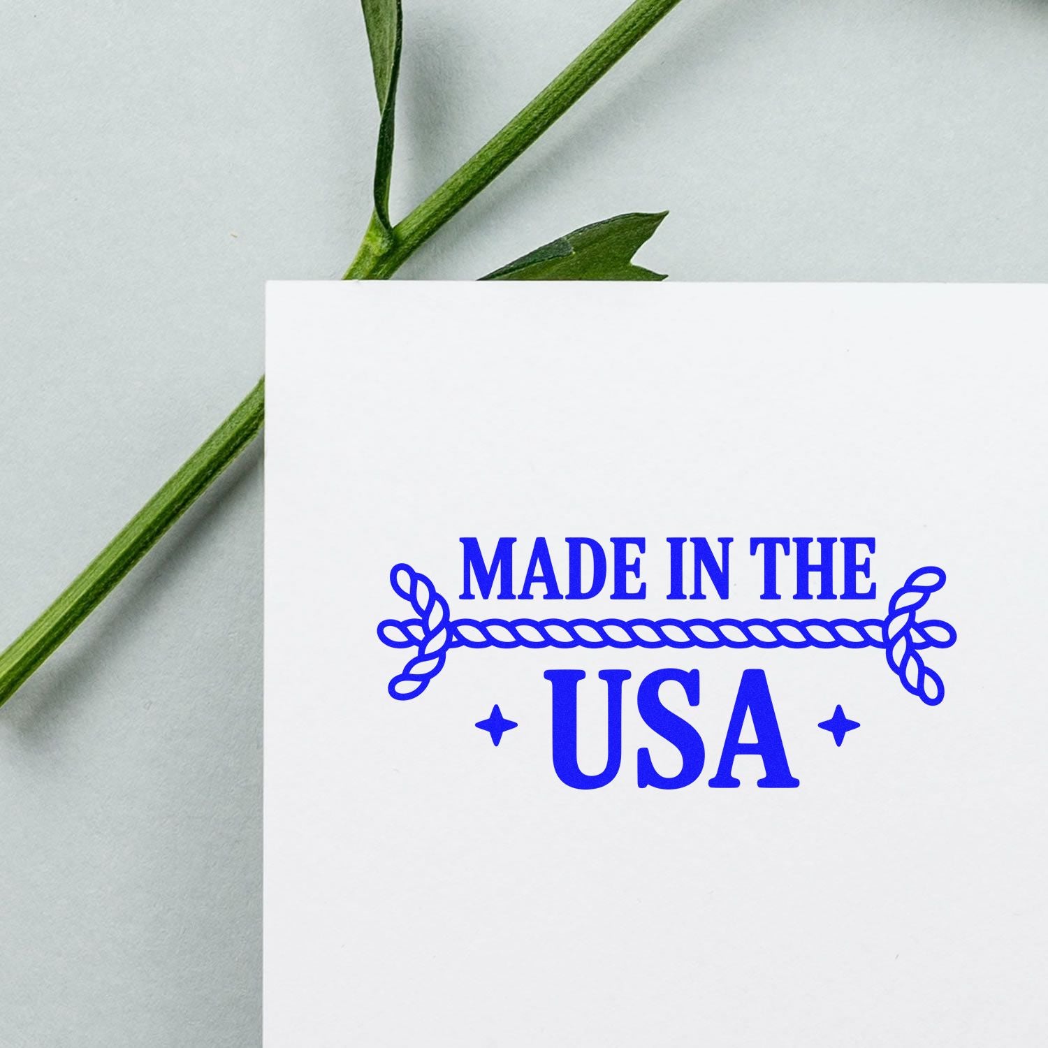 Blue Made in the USA stamp seal with rope border on white card, framed by green stem on a pale background.