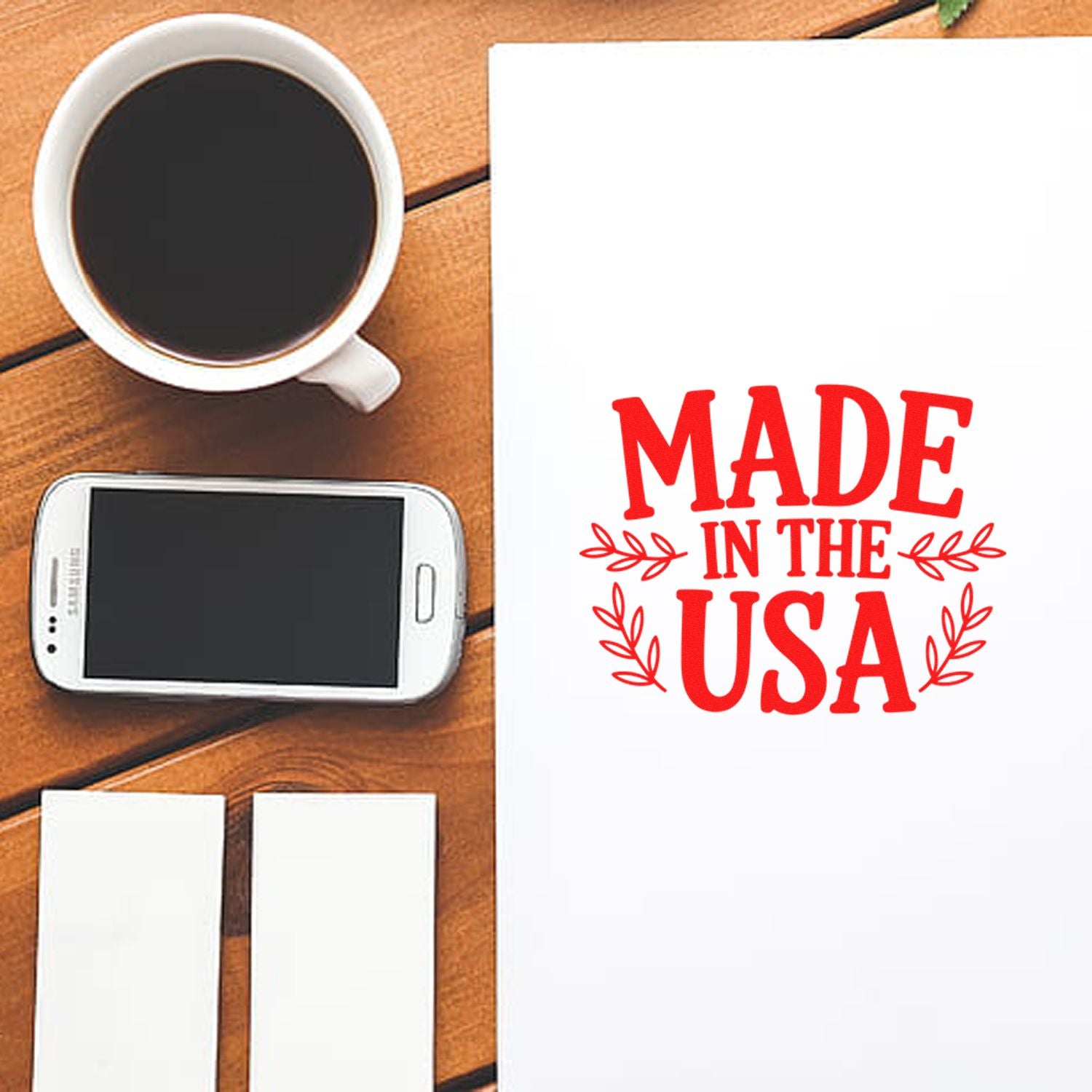 Red Made in the USA stamp and seal set on white paper, beside a black coffee mug and smartphone on a wood desk.