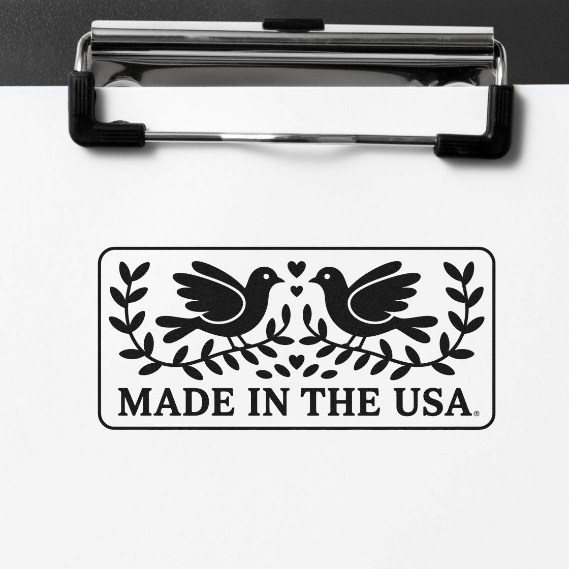 Black Made in the USA stamp seal with two birds, leaves and hearts on white paper, held by a silver clipboard.