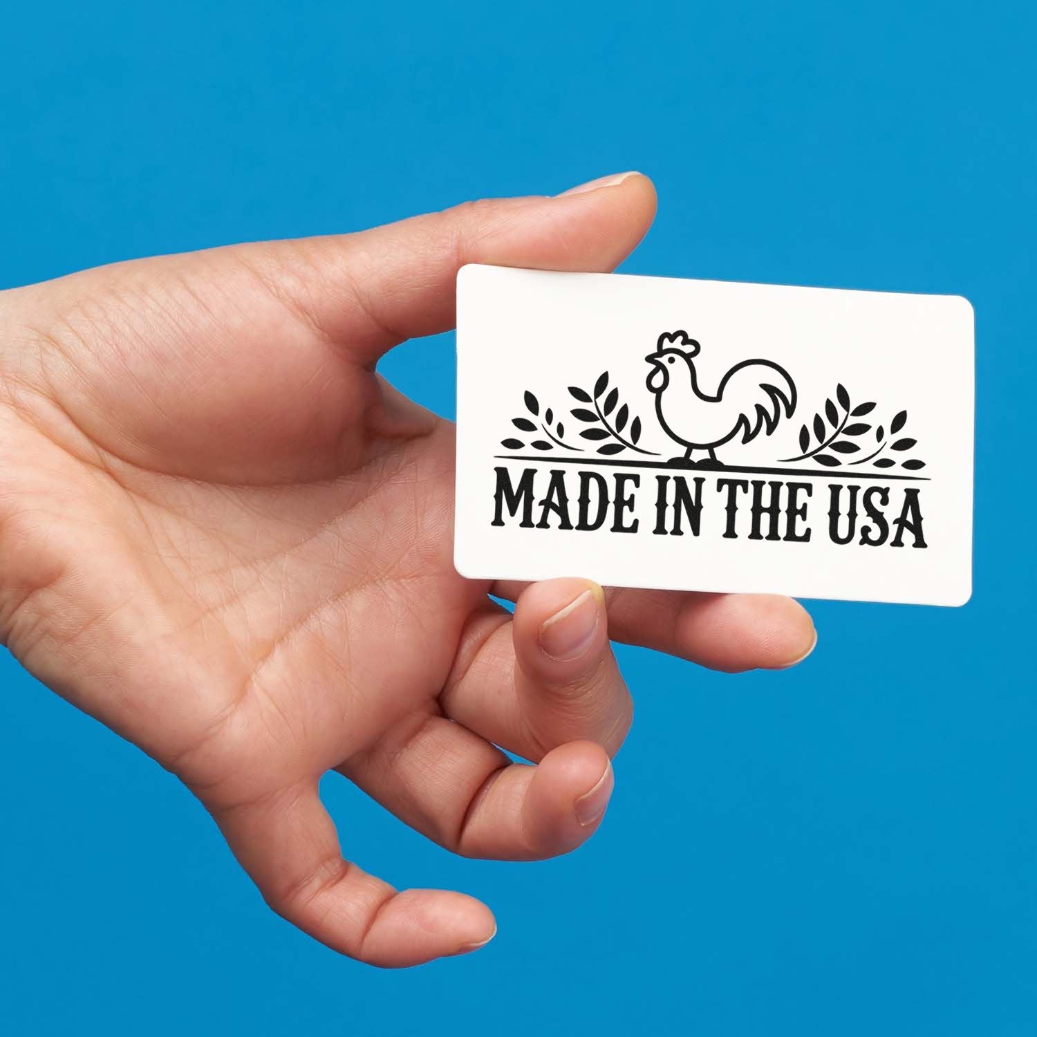 Hand holding white Made in the USA stamp and seal card with black rooster, leaves, and bold black text on blue background.