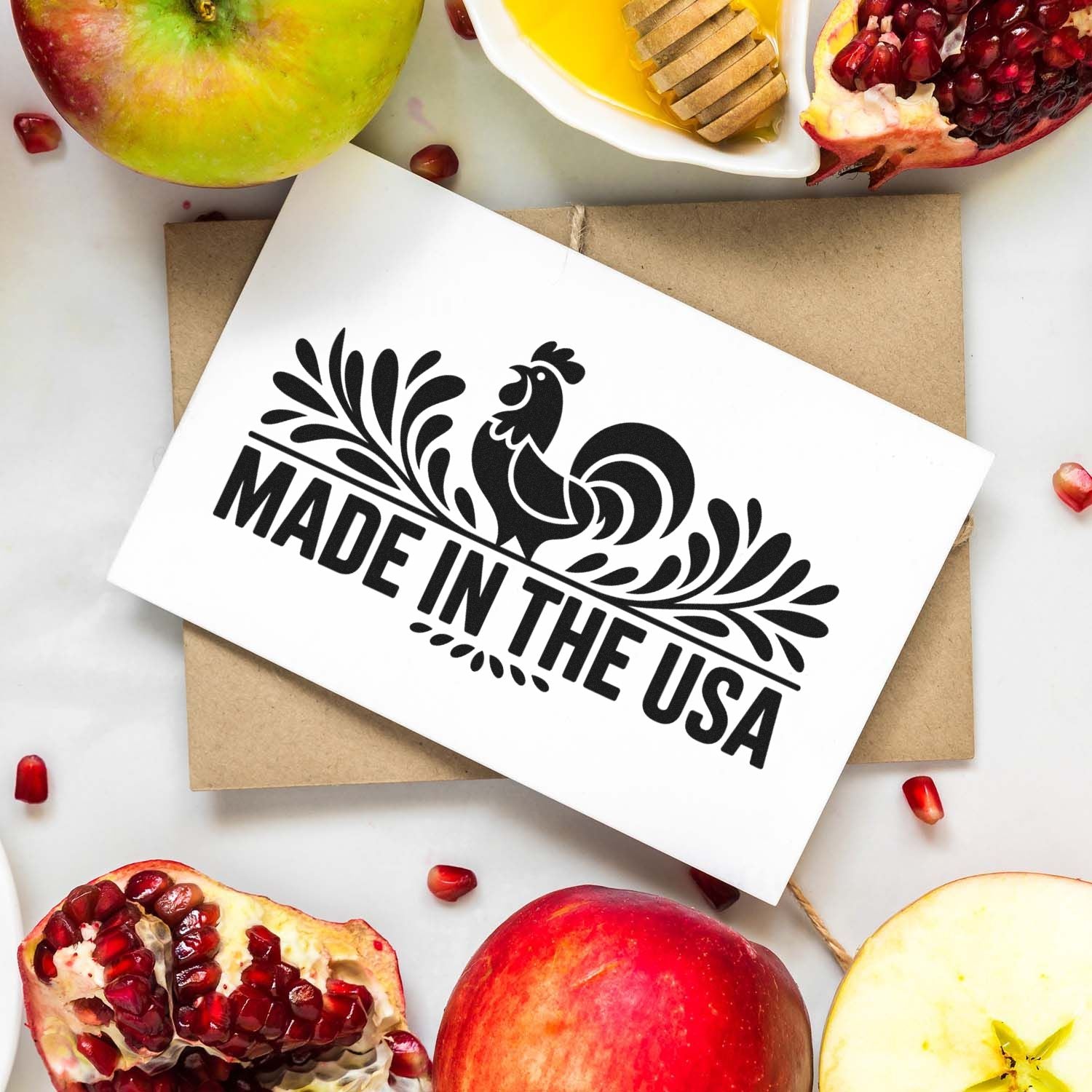 Custom stamp and seal with black rooster and Made in the USA text on white card, over kraft envelope with apples and pomegranates.