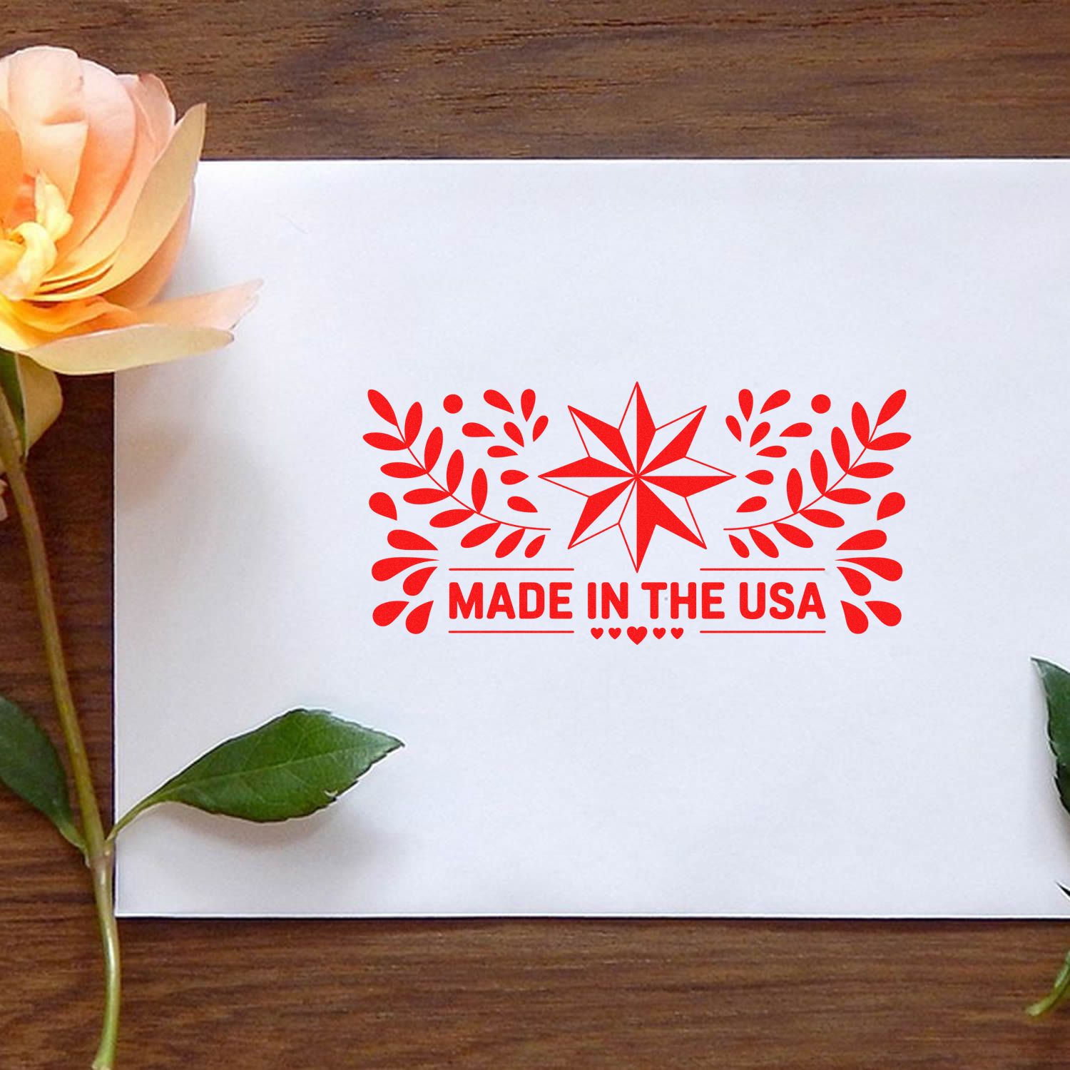 Red Made in the USA stamp seal with floral star design on white card, set on a wood table with flowers and leaves.