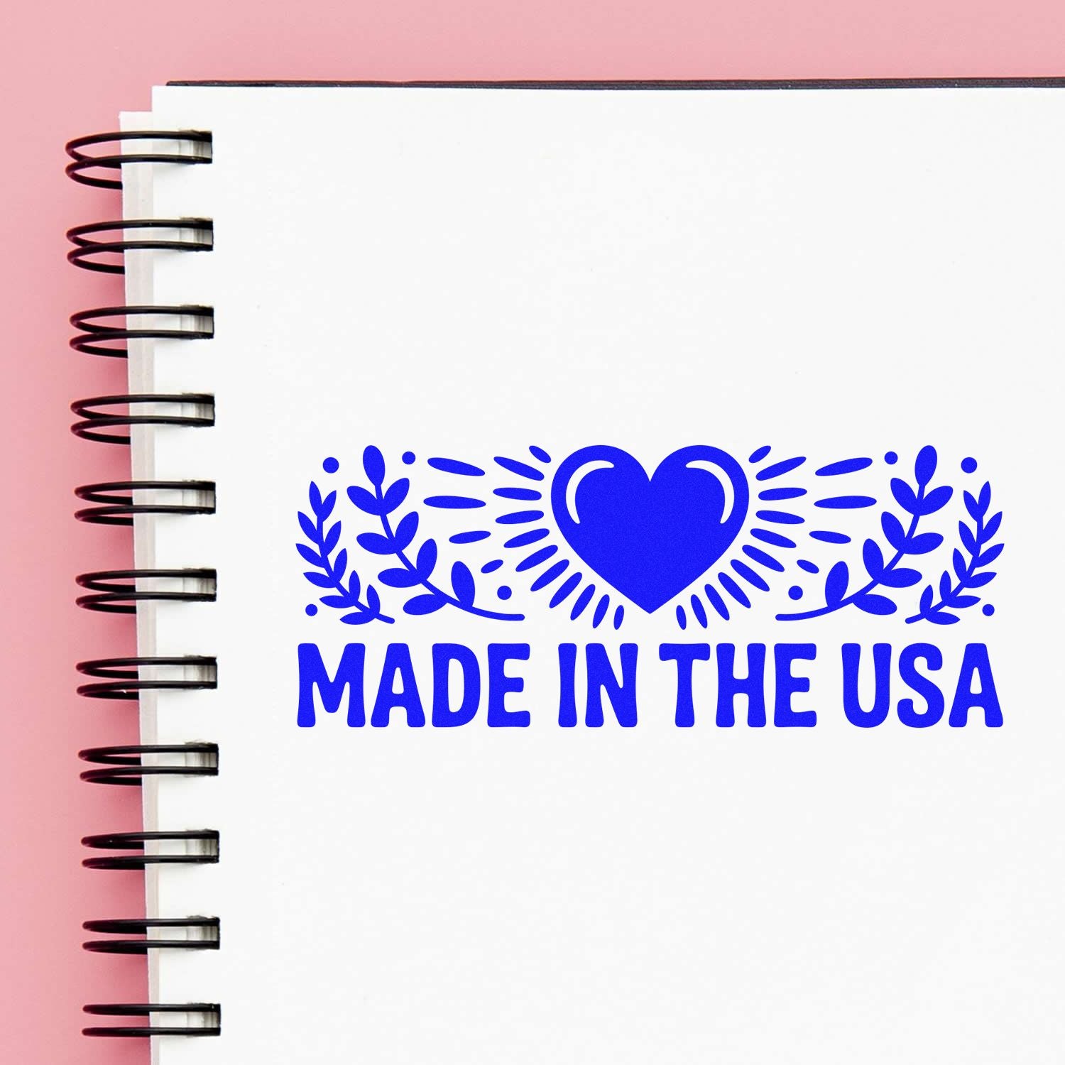 Blue Made in the USA stamp and seal with heart and laurel on a white spiral notebook, set against a pink background.